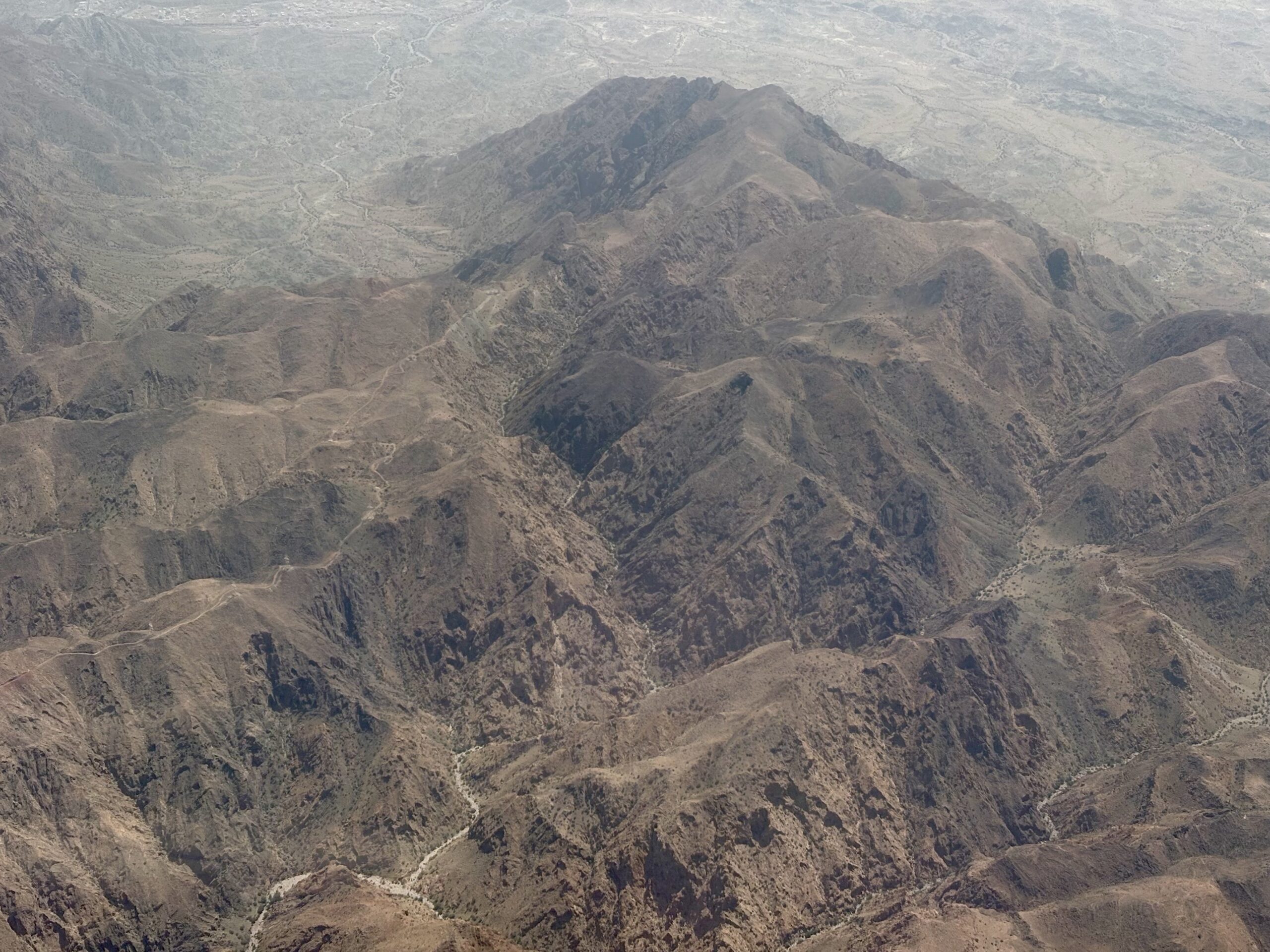 Aerial view of rugged brown mountains and dry valleys with winding trails and sparse vegetation in a desert landscape.