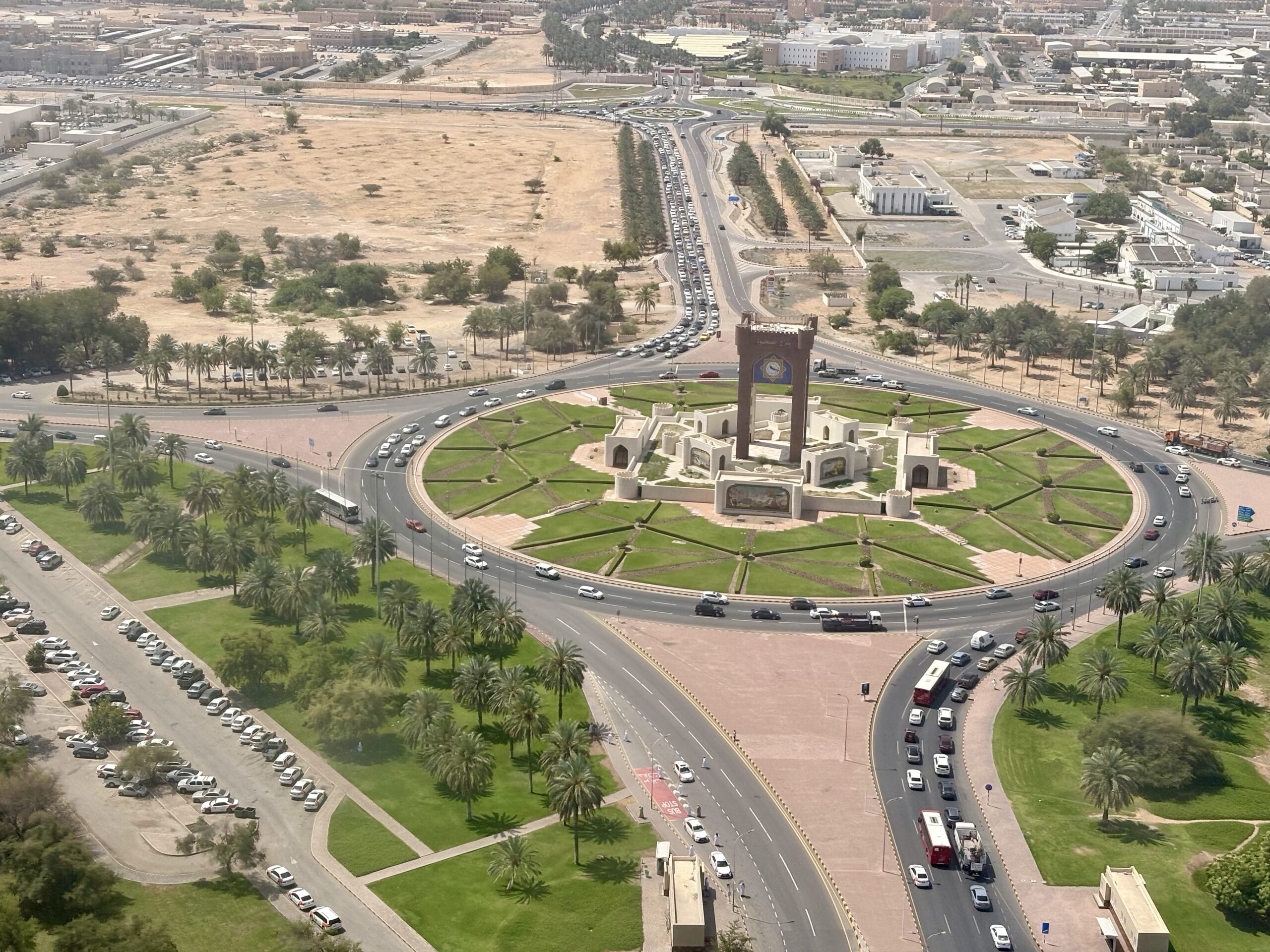 Aerial view of a large roundabout with a tall brick monument at its center, green lawns, and palm trees, with a long line of cars on the surrounding road.