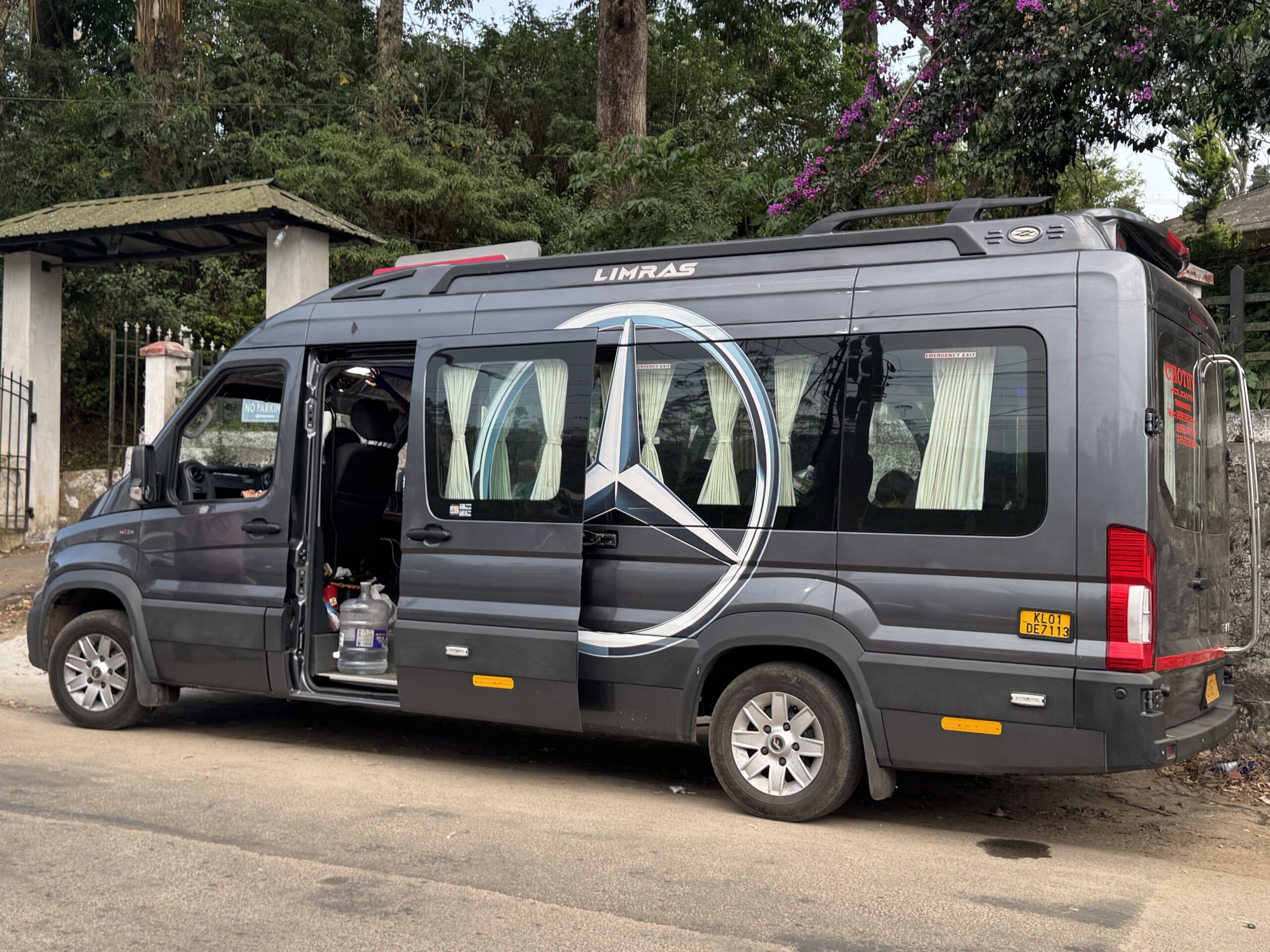 Dark gray van with open side door, showing cushioned seats and light curtains in the passenger windows, parked on a street with greenery in the background.