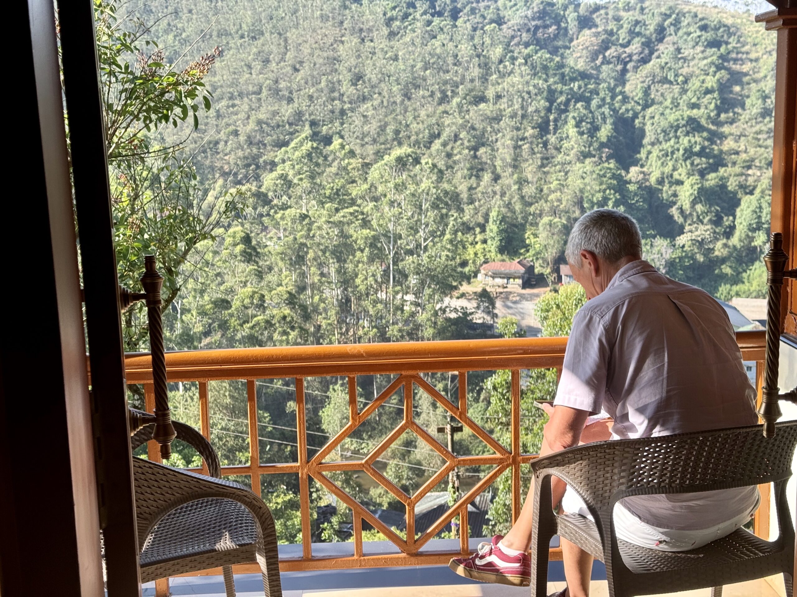 Man in a light shirt seated on a wicker chair on a balcony, facing a lush forested hillside.
