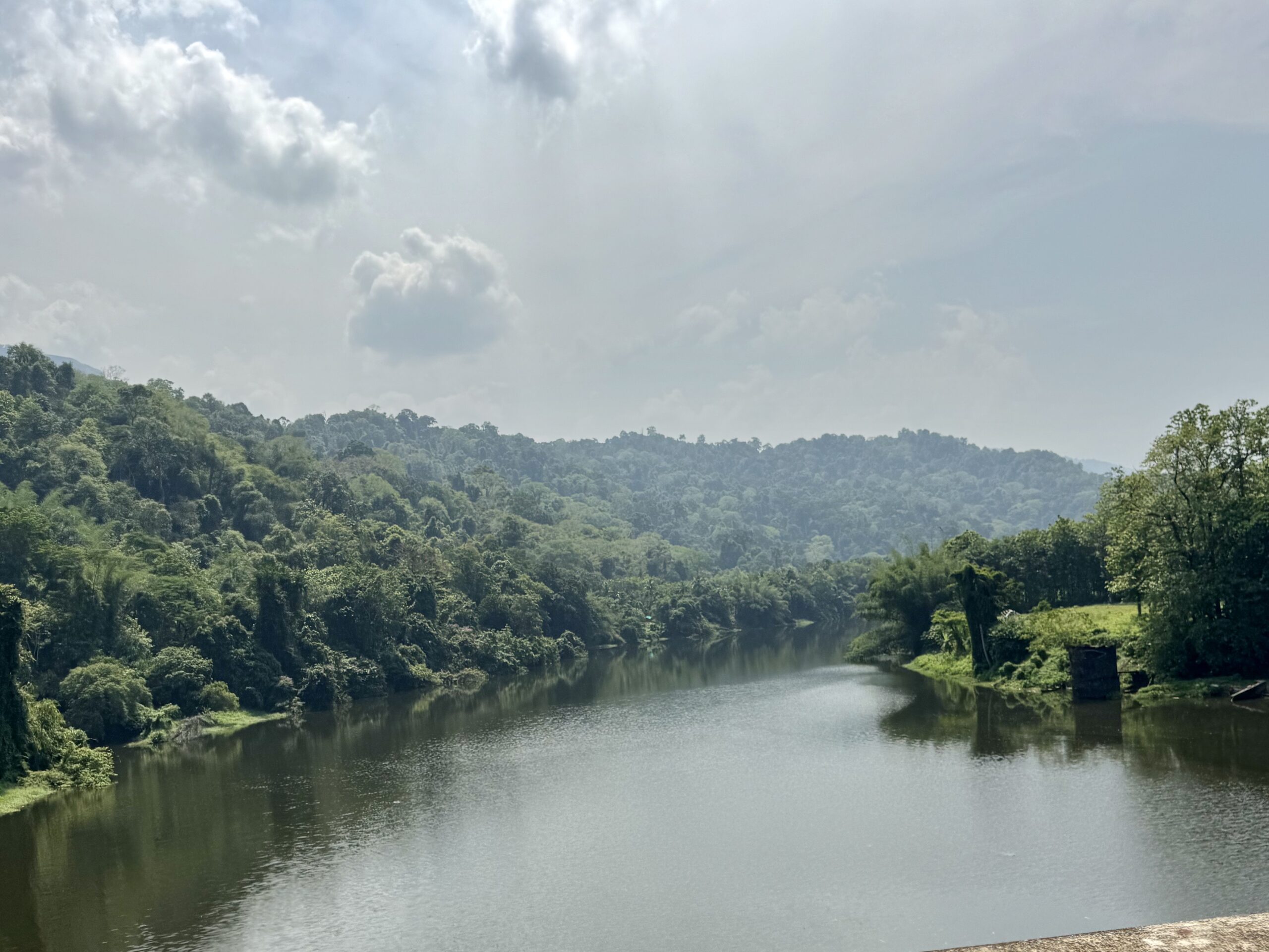 River winds through green forested hills with a calm, reflective body of water beneath a partly cloudy sky.