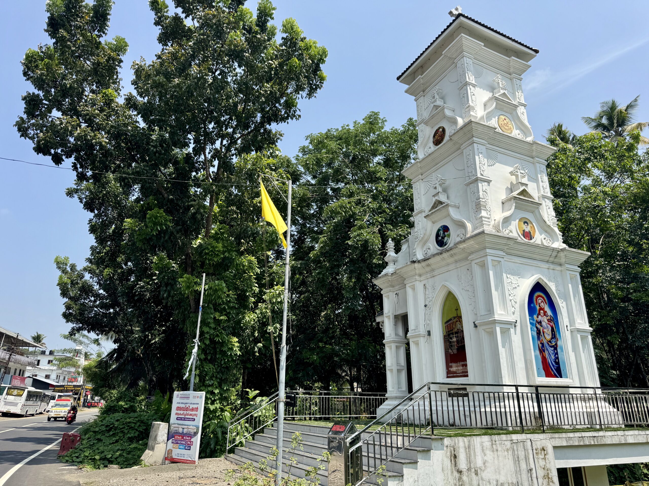 White ornate church tower with stairs and railing on a sunny street, flanked by tall green trees and a blue sky.