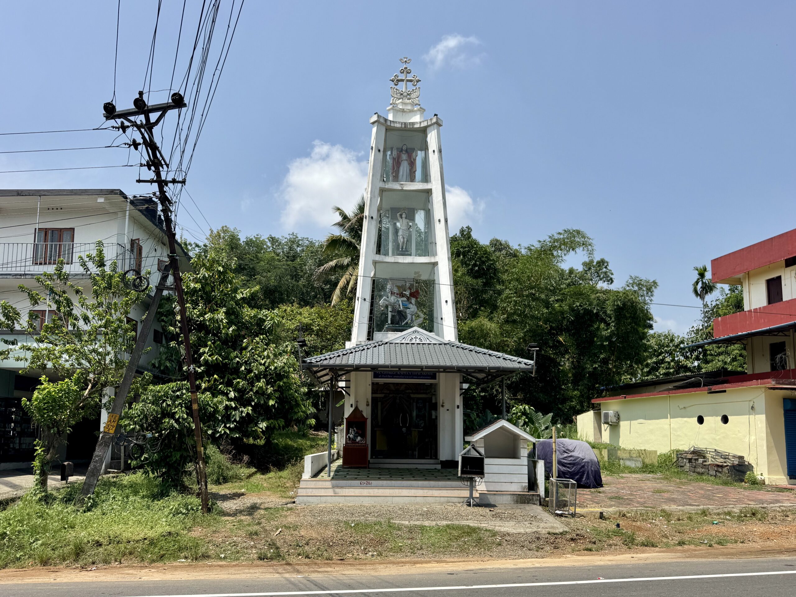 White shrine tower with glass panels showing statues, topped by a cross, in a sunny urban setting.