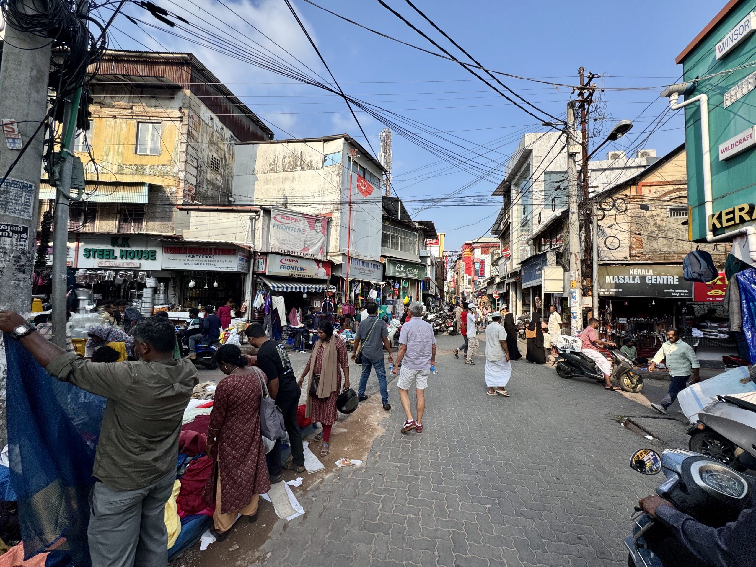 Busy outdoor market street in India with stalls, shoppers, and tangled overhead wires.