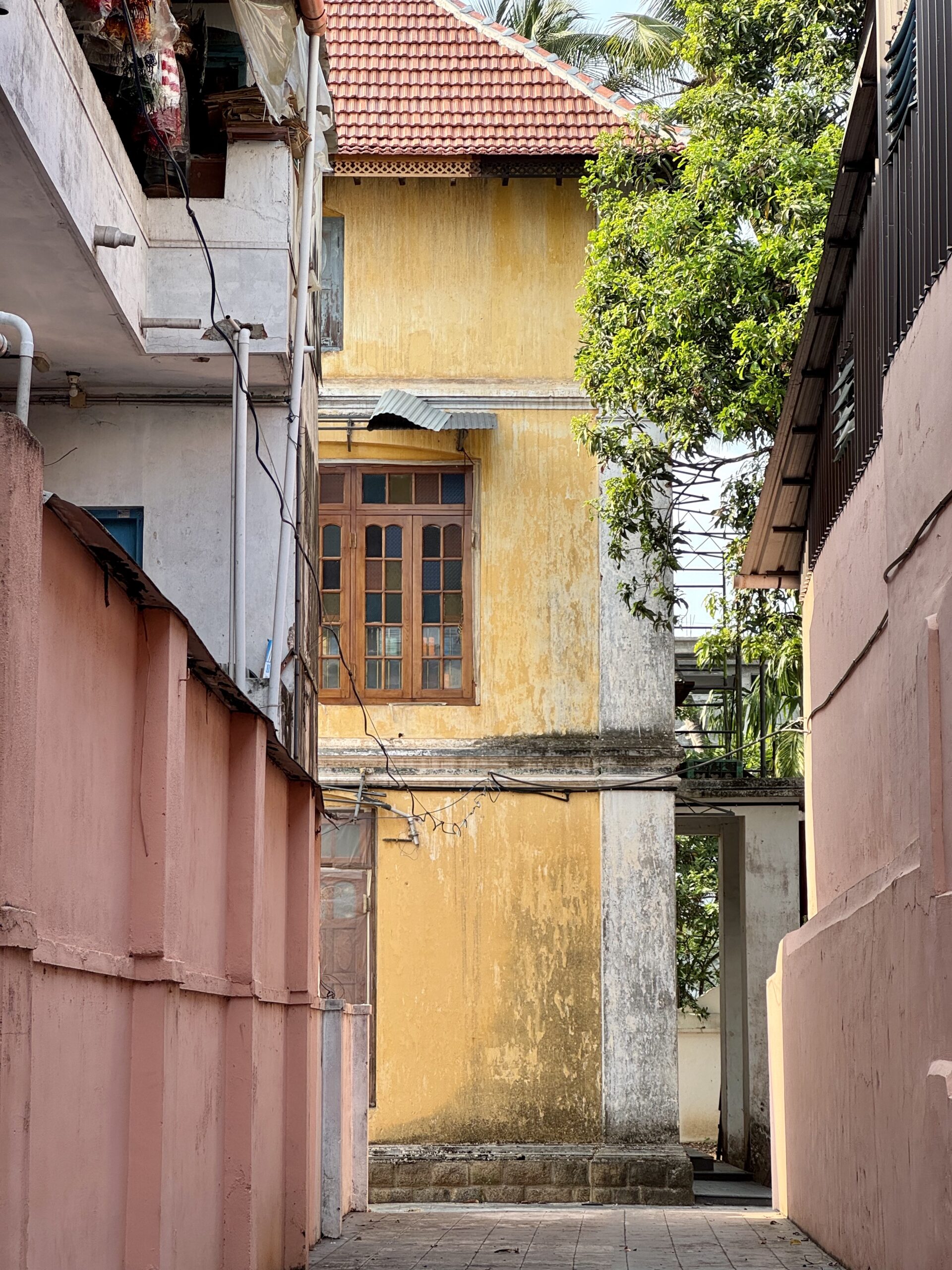 Narrow sunlit alley between pink walls, revealing a yellow building with a wooden window and a small metal awning.