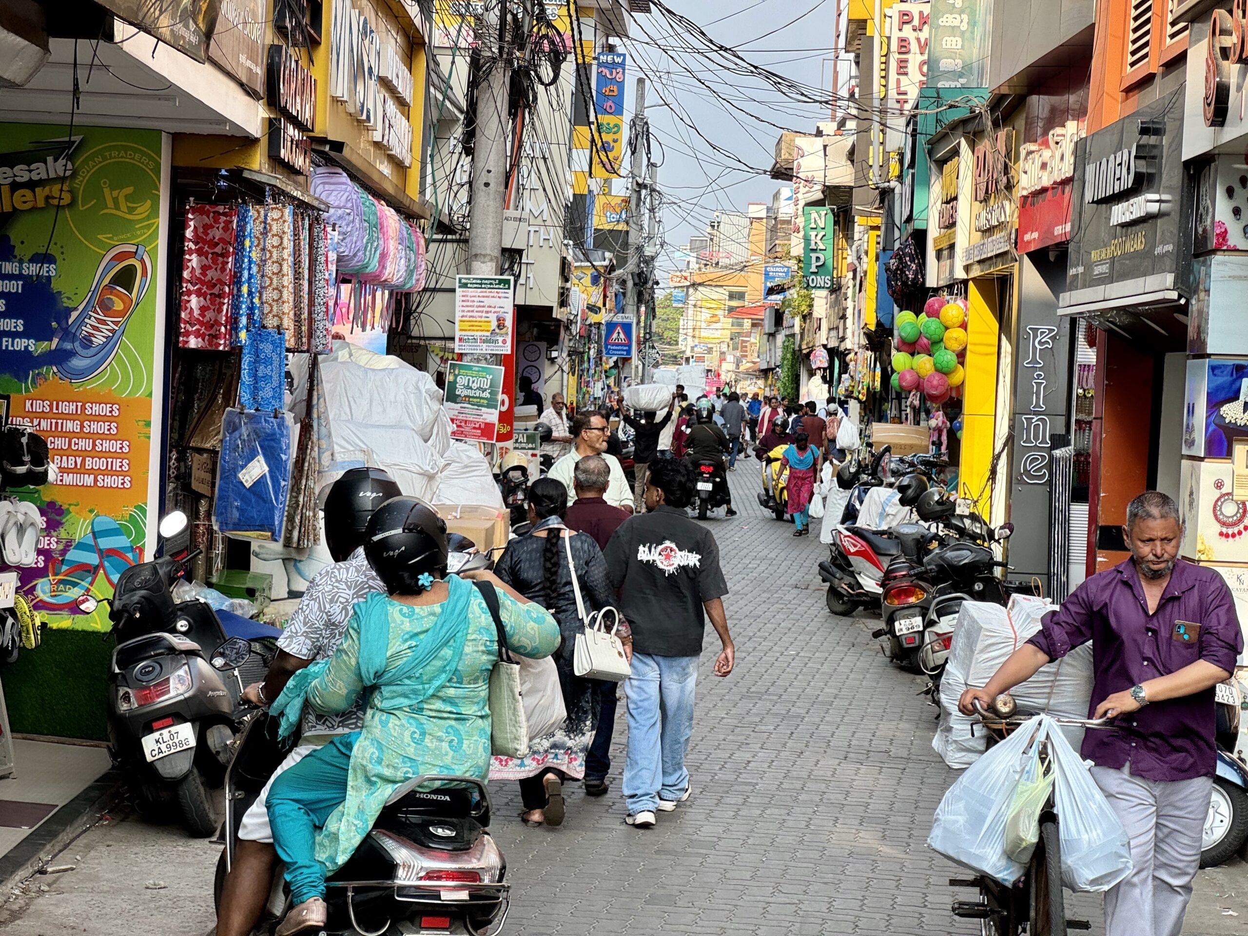 Busy street market in India with scooters parked along the sides and colorful shopfronts on both sides.