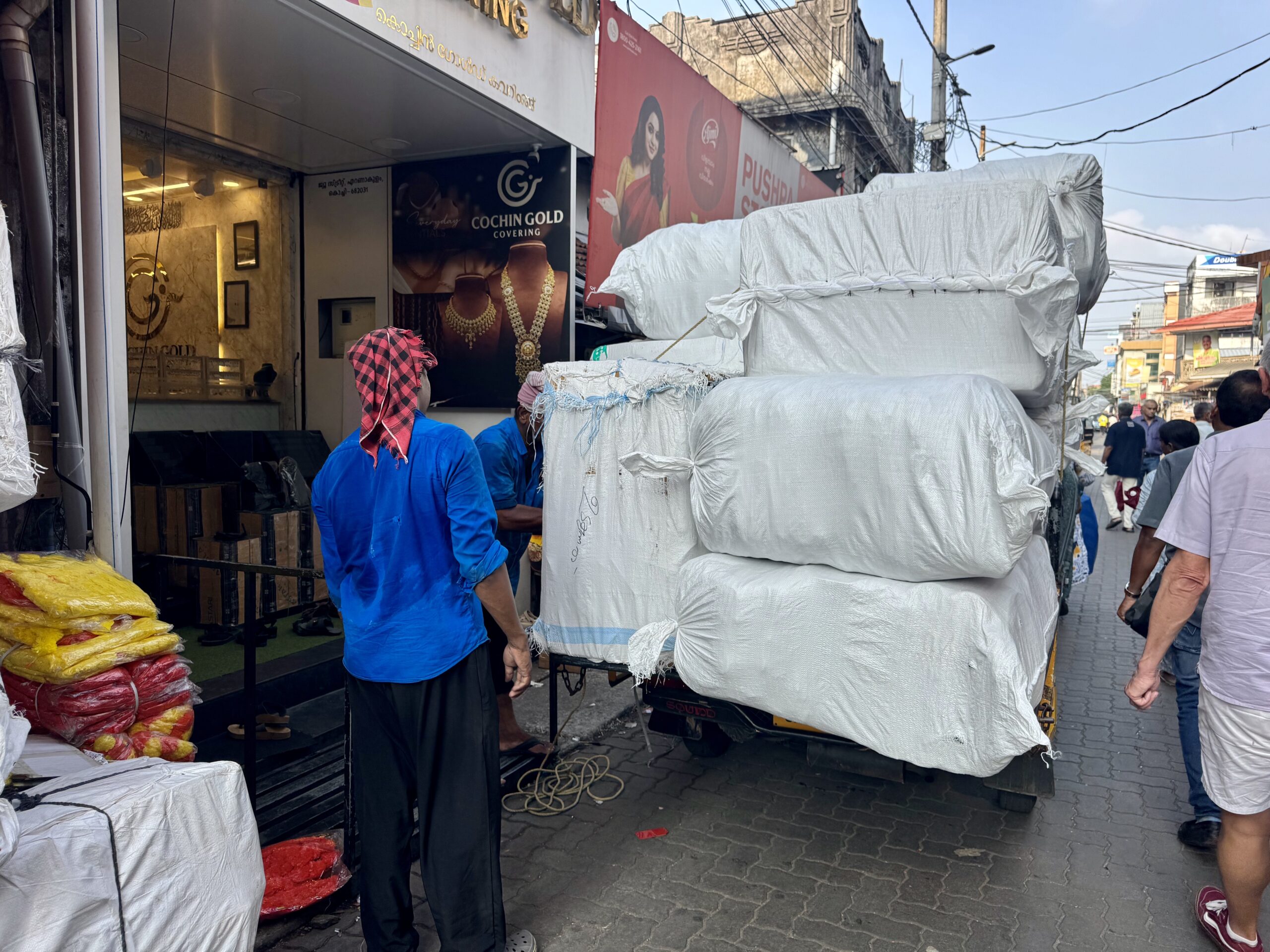 Men in blue uniforms stand near a white-wrapped cargo stack on a truck in a busy street with a jewelry shop facade nearby.
