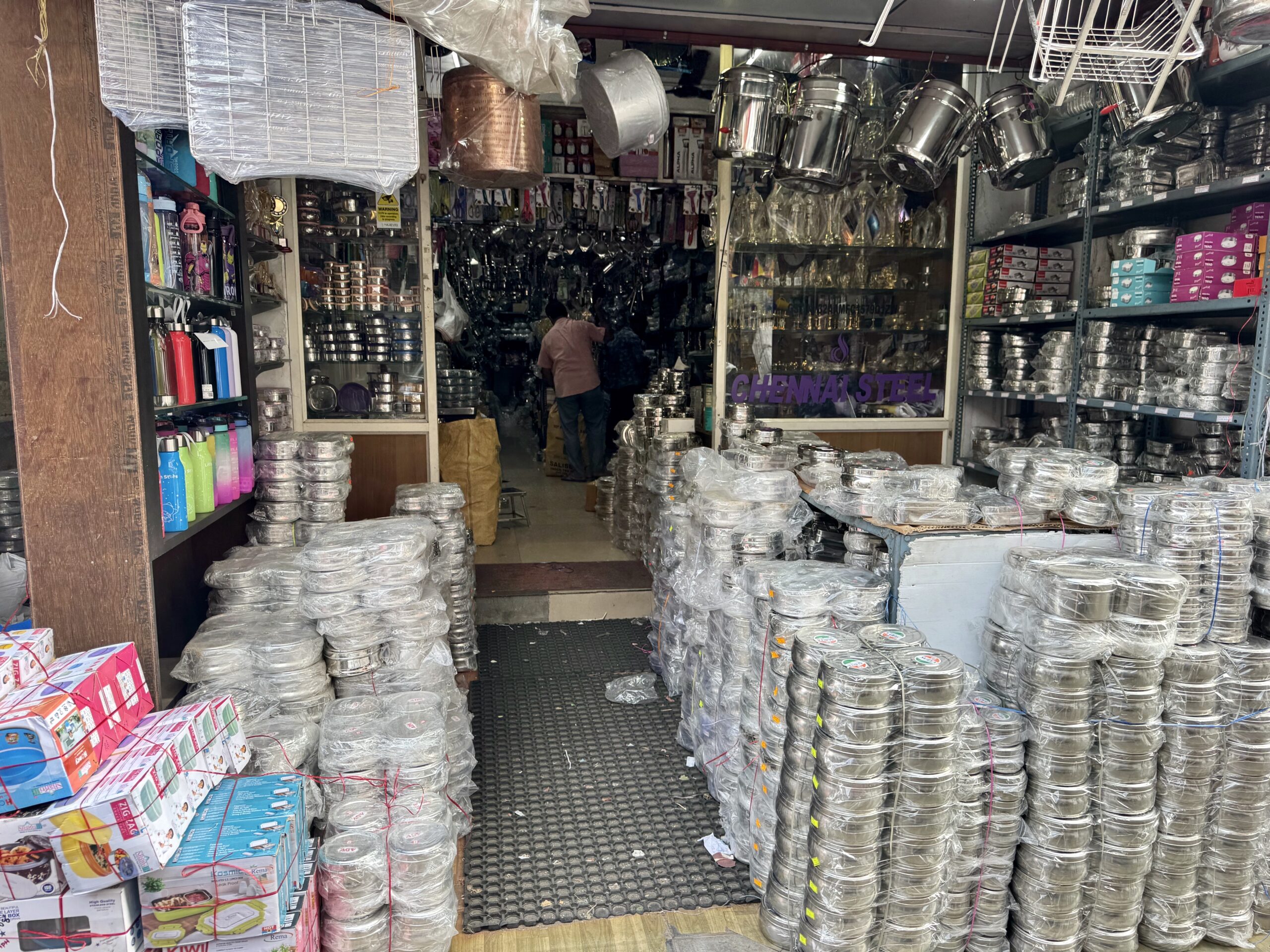 Narrow cookware shop filled with stacked aluminum pots and pans, shelves on both sides and customers in the back line of sight, center.
