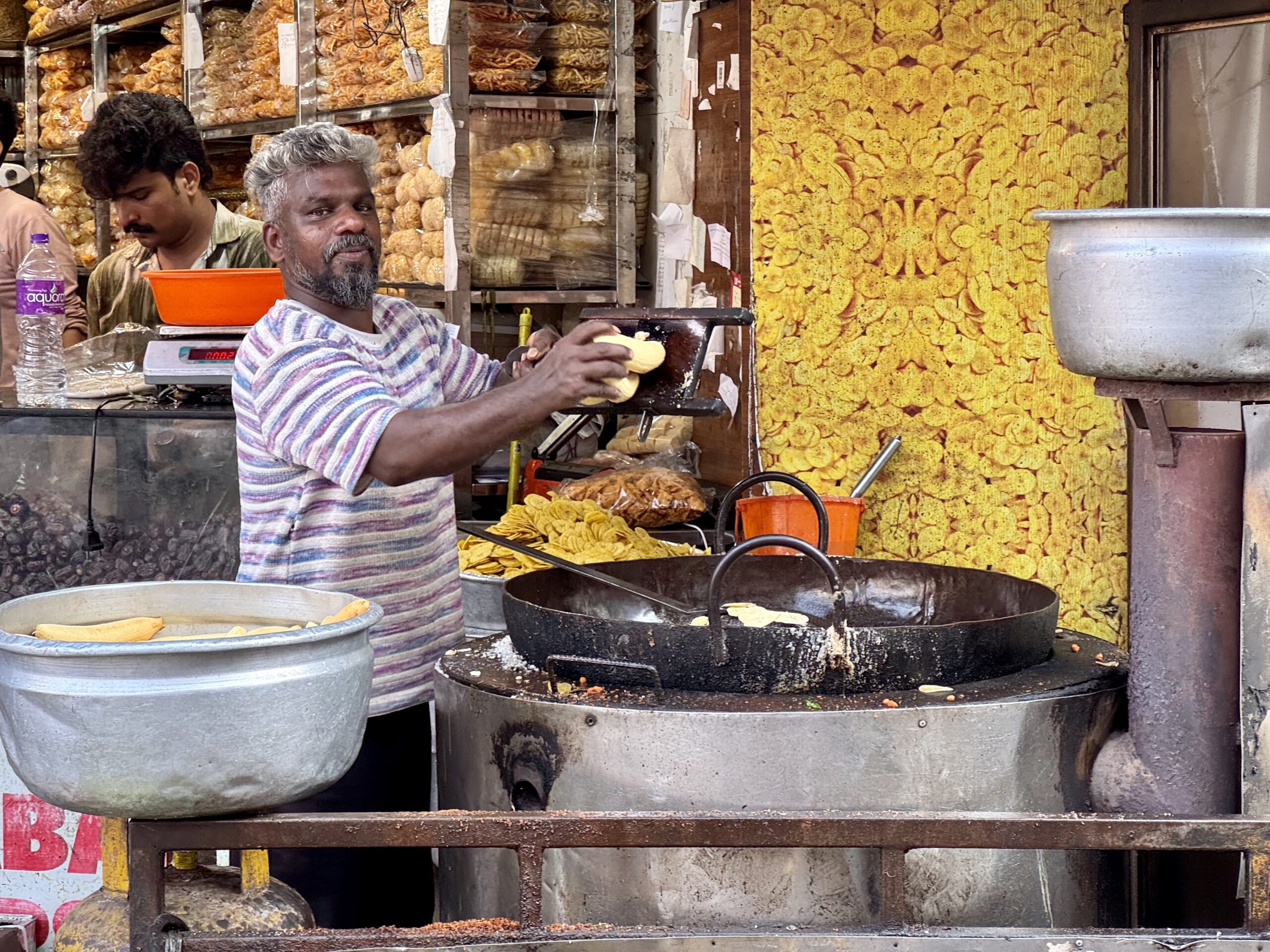 Street food vendor frying round yellow snacks in a large pan, with shelves of chips behind him