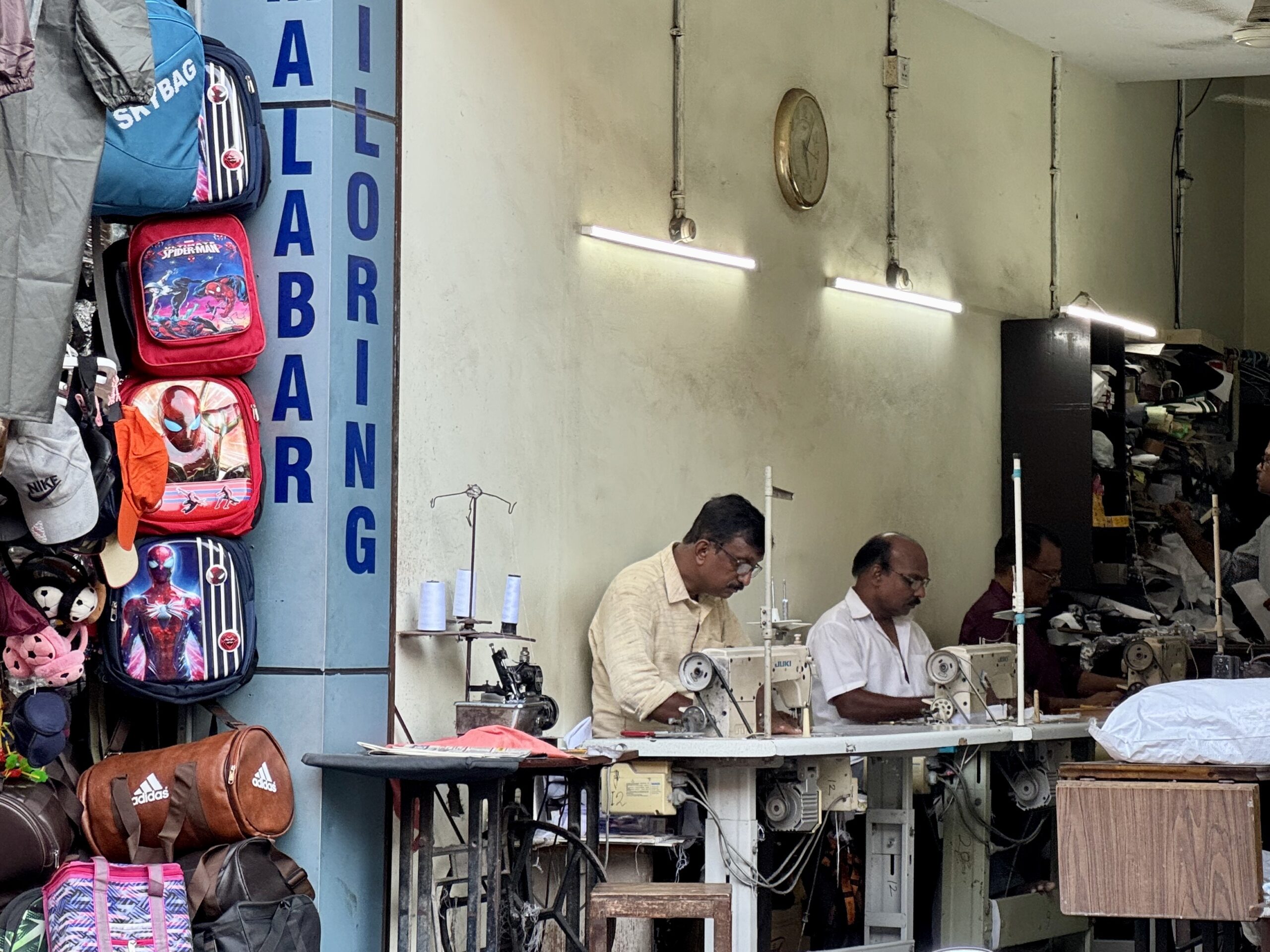Tailors at work in a busy workshop with sewing machines along a worktable, backpacks hanging on the left wall and storage on the right side.