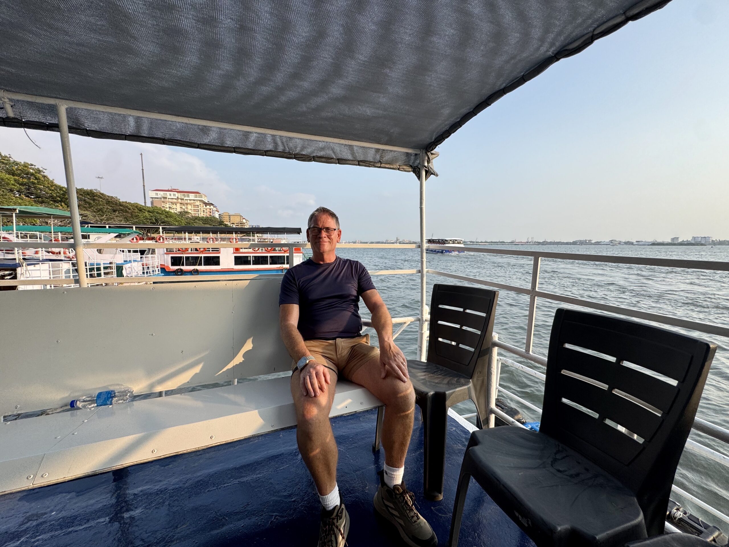 Man seated on a boat deck under a shade sail, with water, chairs, and distant buildings visible in the background.