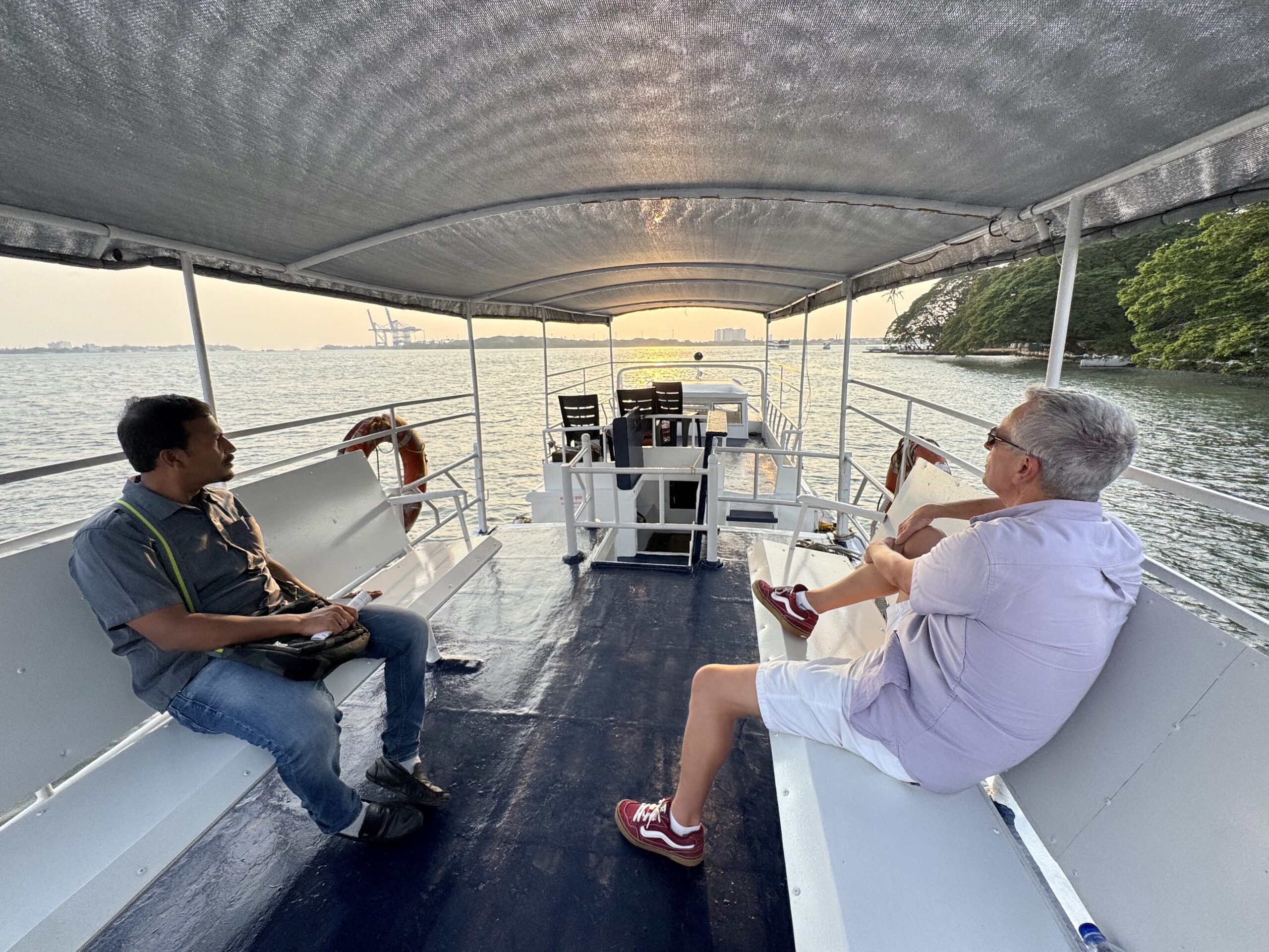 Two men sit on a covered boat deck facing aft with water and a distant shoreline in view during sunset.