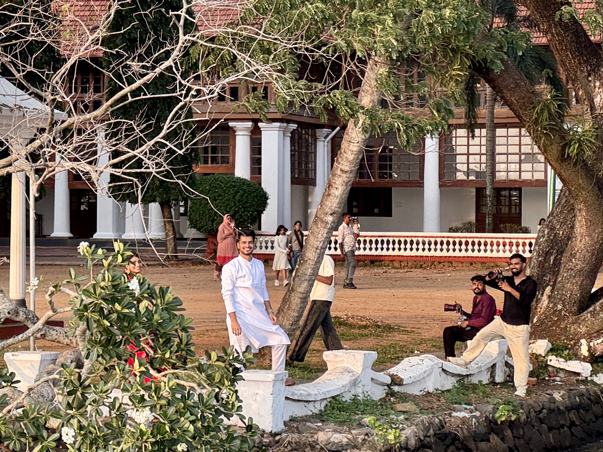 Group of people in traditional attire outside a colonial-style building with white columns, trees, and shrubbery.