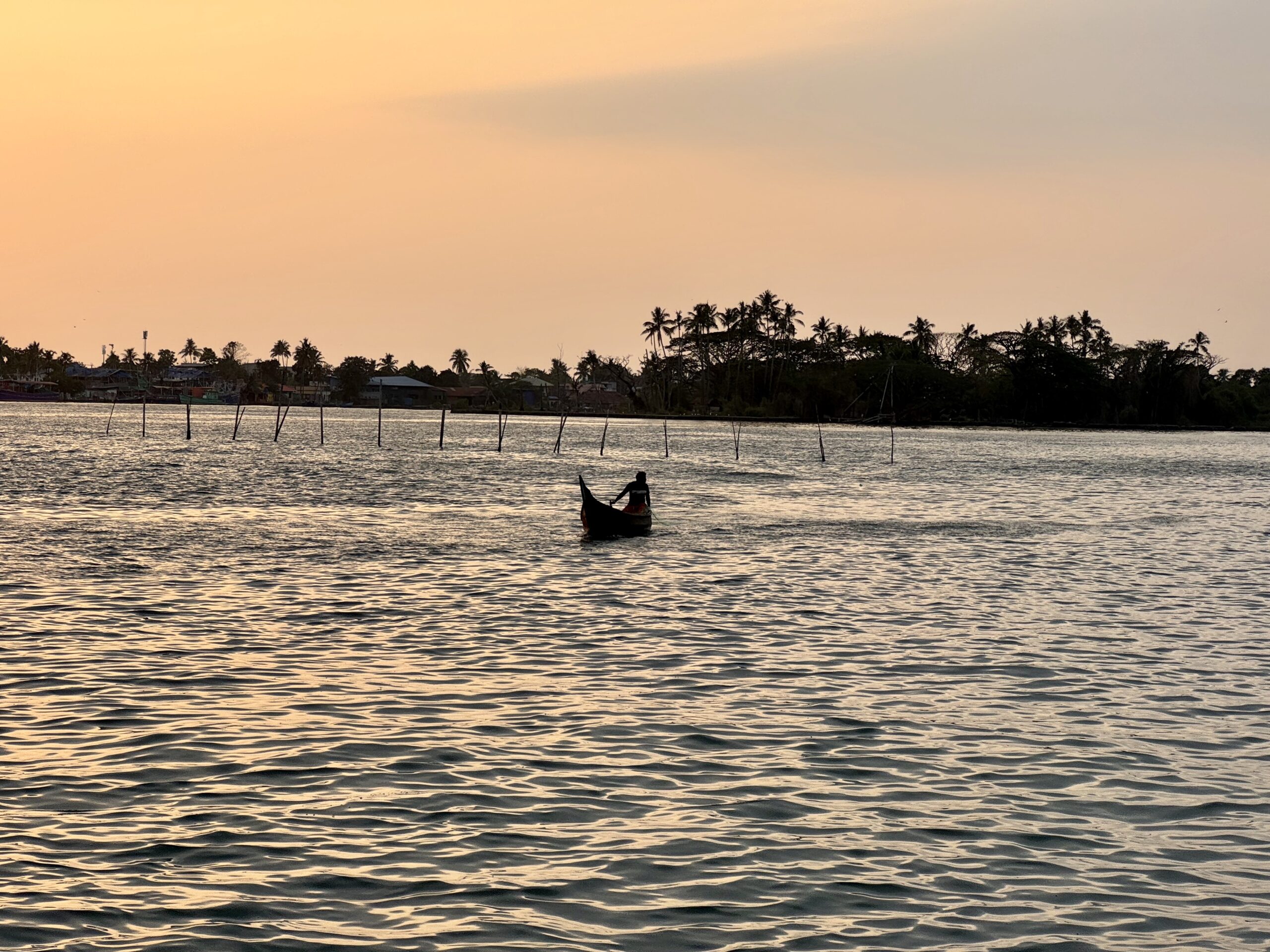 Silhouetted boat on calm water at sunset with a palm-tree shoreline in the distance.