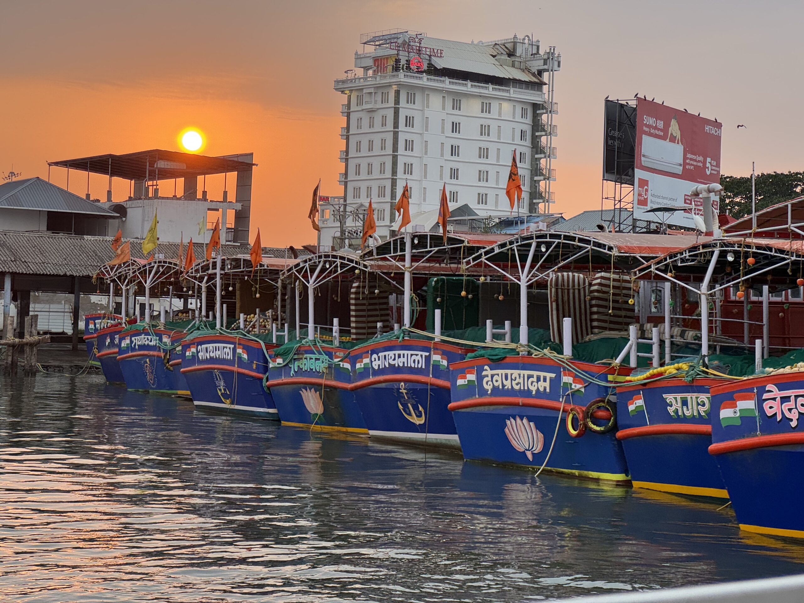 Row of colorful fishing boats tied at a dock at sunset, with an orange sky and calm water reflecting hues.