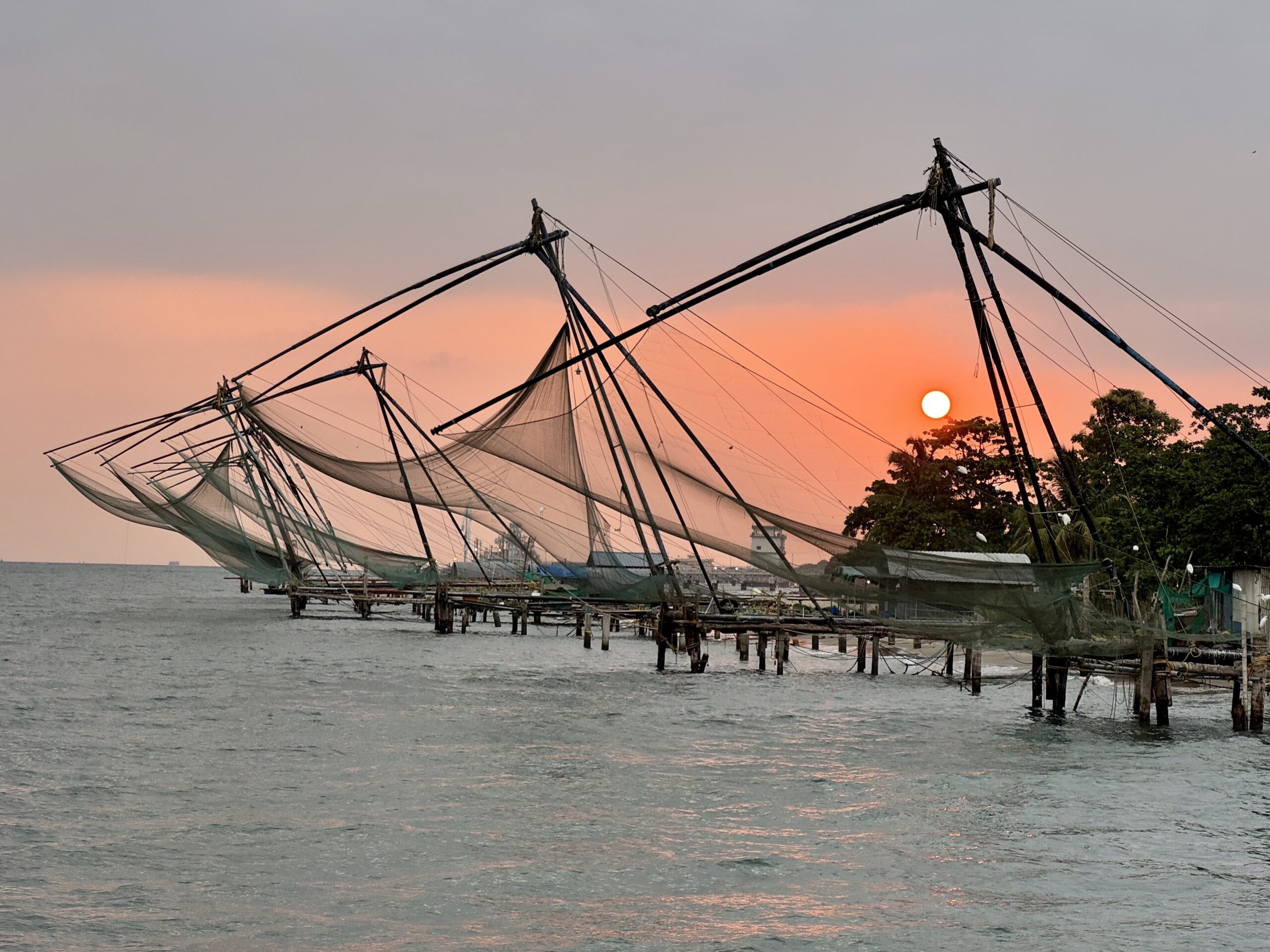 Sunset over a fishery with large nets stretched between tall poles over calm water near a shoreline.