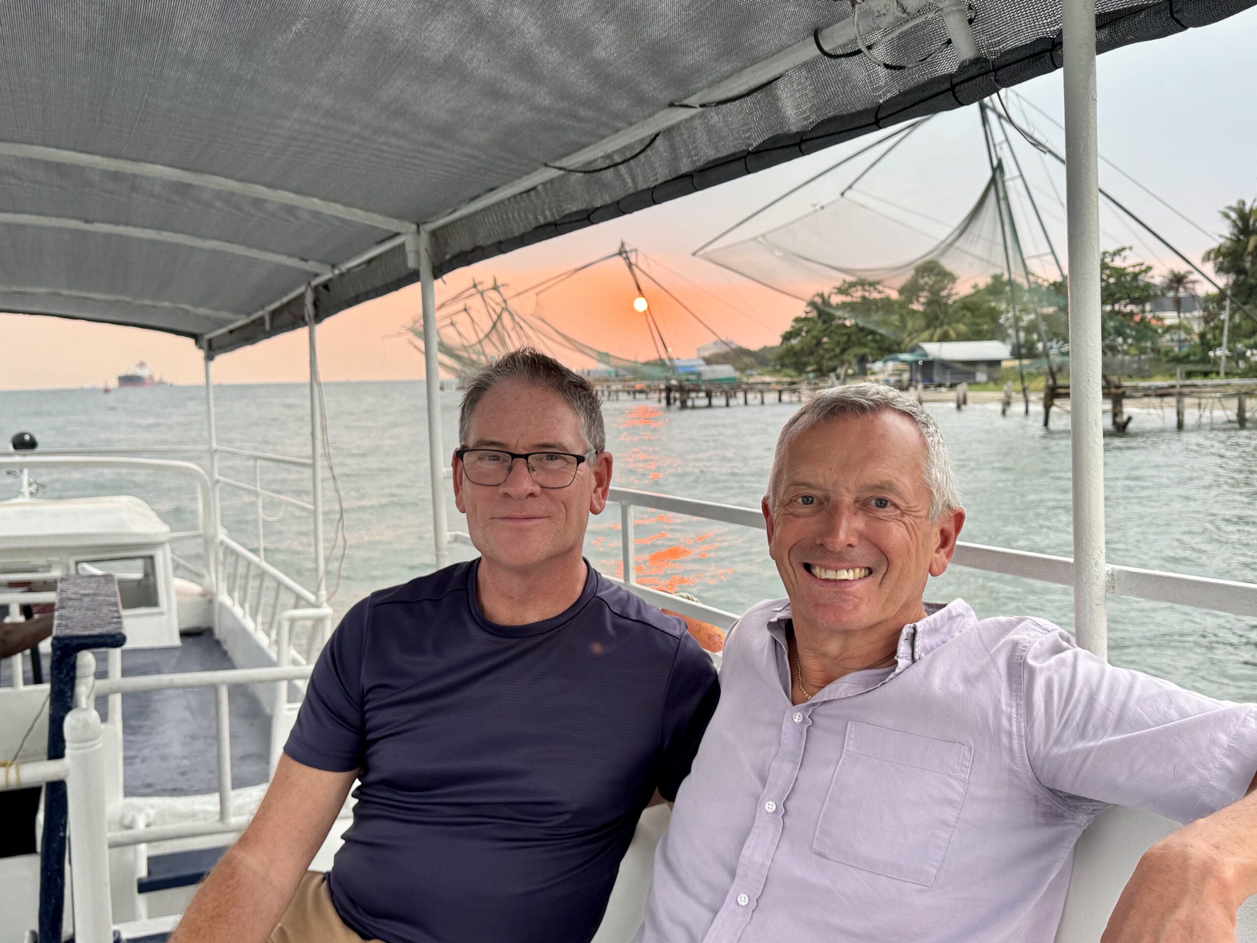 Two men posing for a selfie on a boat deck at sunset with water and shoreline in the background.
