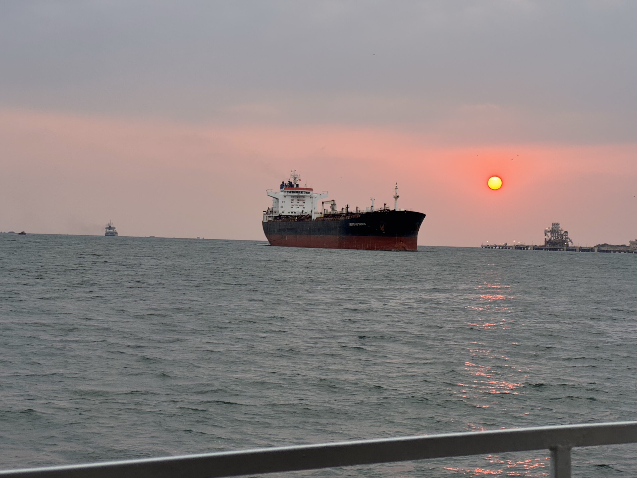 Large cargo ship on a calm sea at sunset, with a railing in the foreground and a pink-orange sky behind it