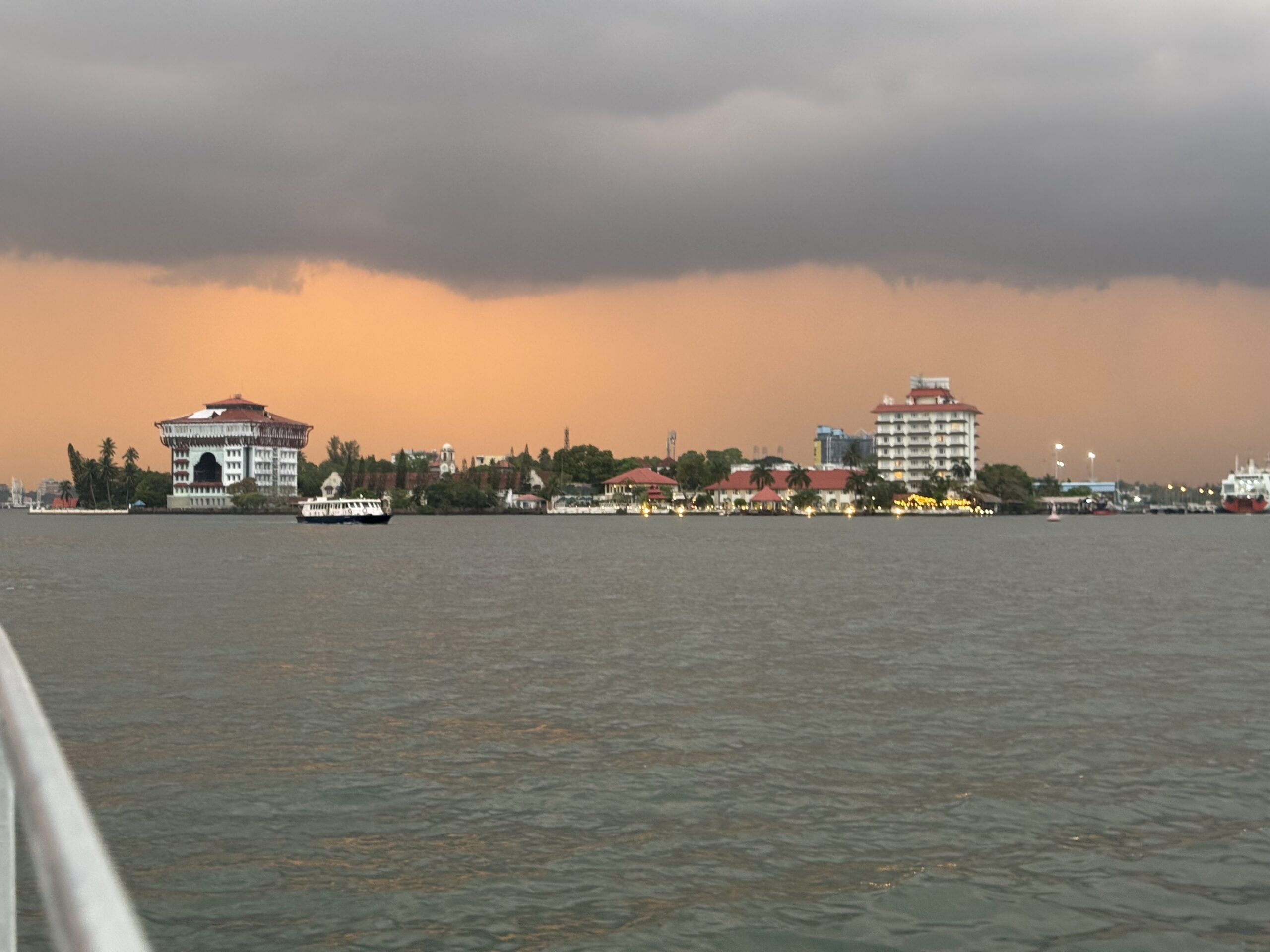 Waterfront city skyline at sunset with orange-hued sky, a white building with a red roof on the left, and a boat on calm water