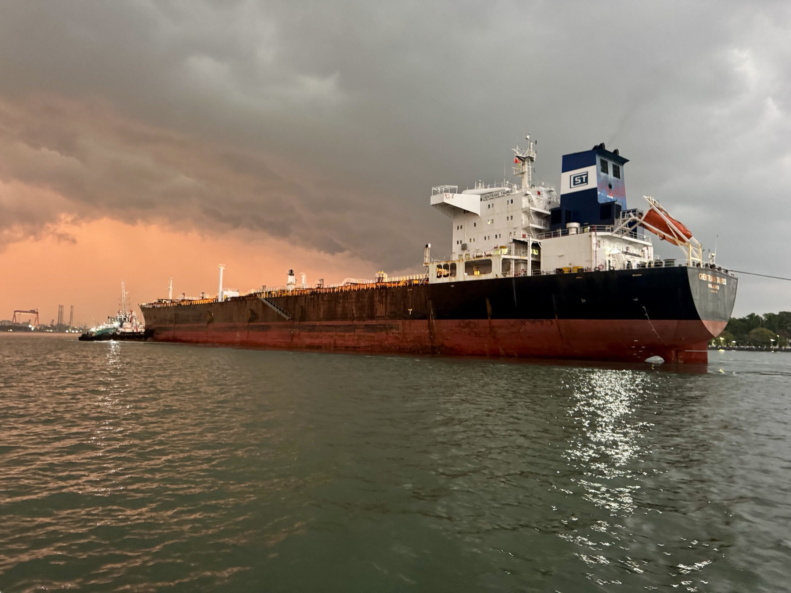Large cargo ship at the port under stormy skies with an orange sunset glow on the water. Important: ship is massive, with a blue control tower visible in the stern area.