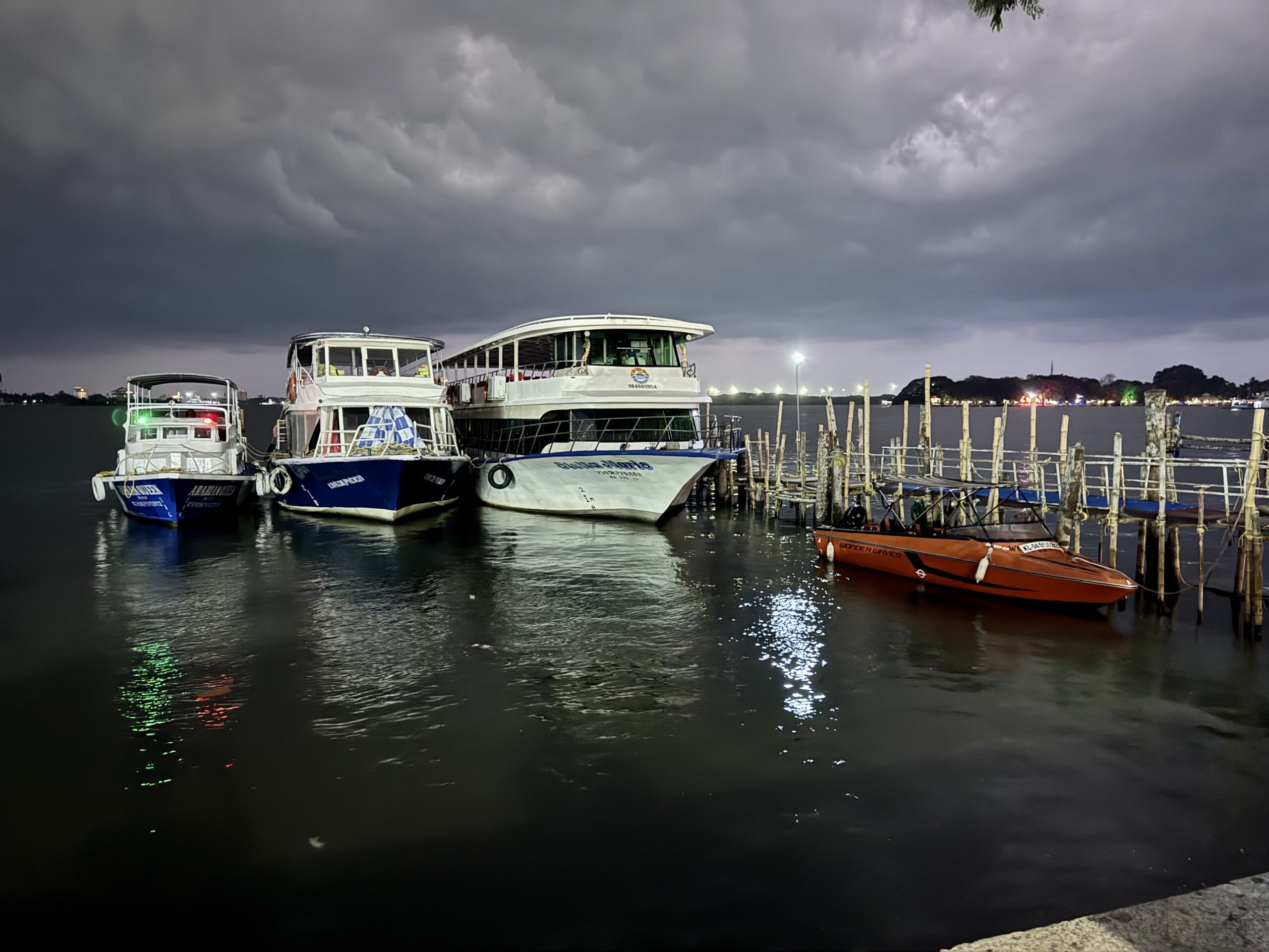 Stormy night over a marina with three white boats docked at a wooden pier and dark clouds overhead, reflections on the water.