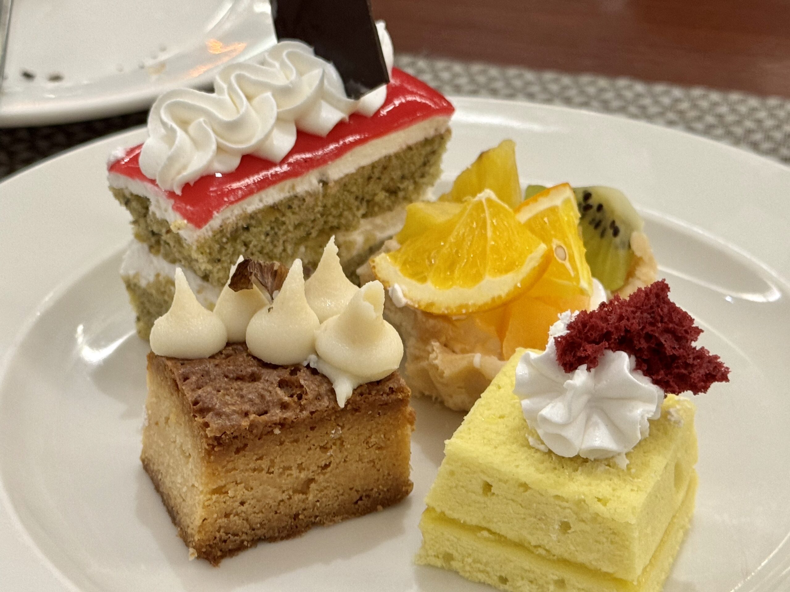 Plate of assorted dessert bites: yellow sponge cake square with whipped cream, orange slices, kiwi, and a chocolate-topped cake piece with white frosting dollops.