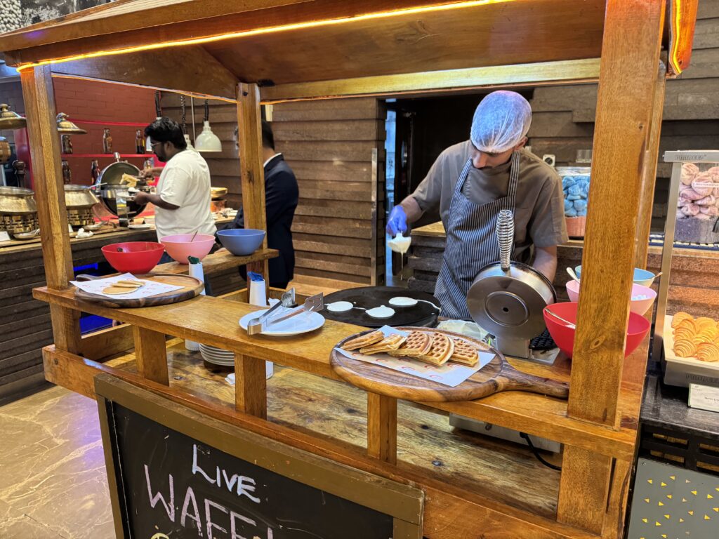Staff behind a wooden market stall making waffles with a hot iron and batter bowls nearby, colorful bowls and toppings visible deep in food prep area.