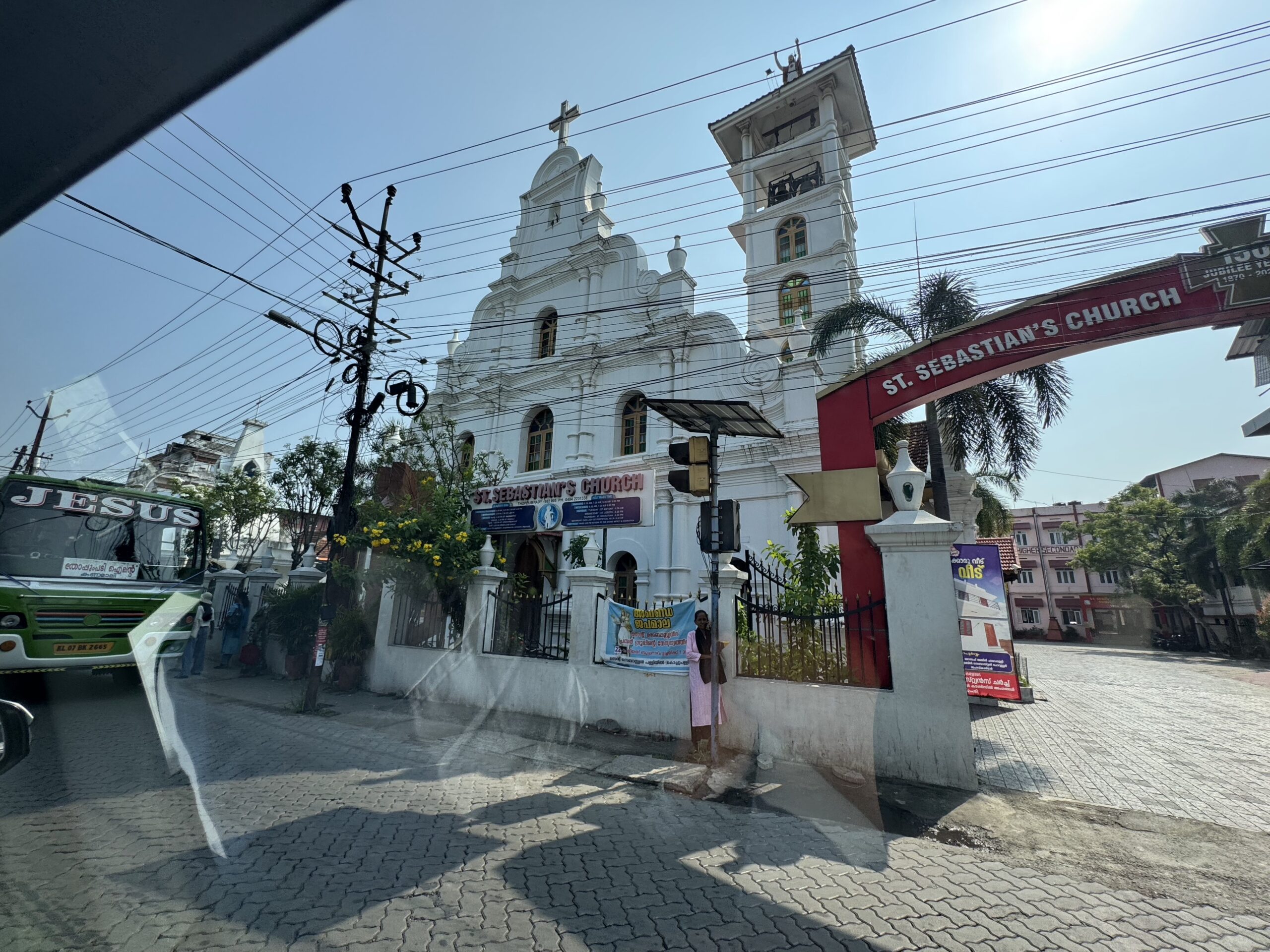 White church with twin bell towers and cross, under a tangle of power lines, near a red arch reading St. Sebastian's Church.