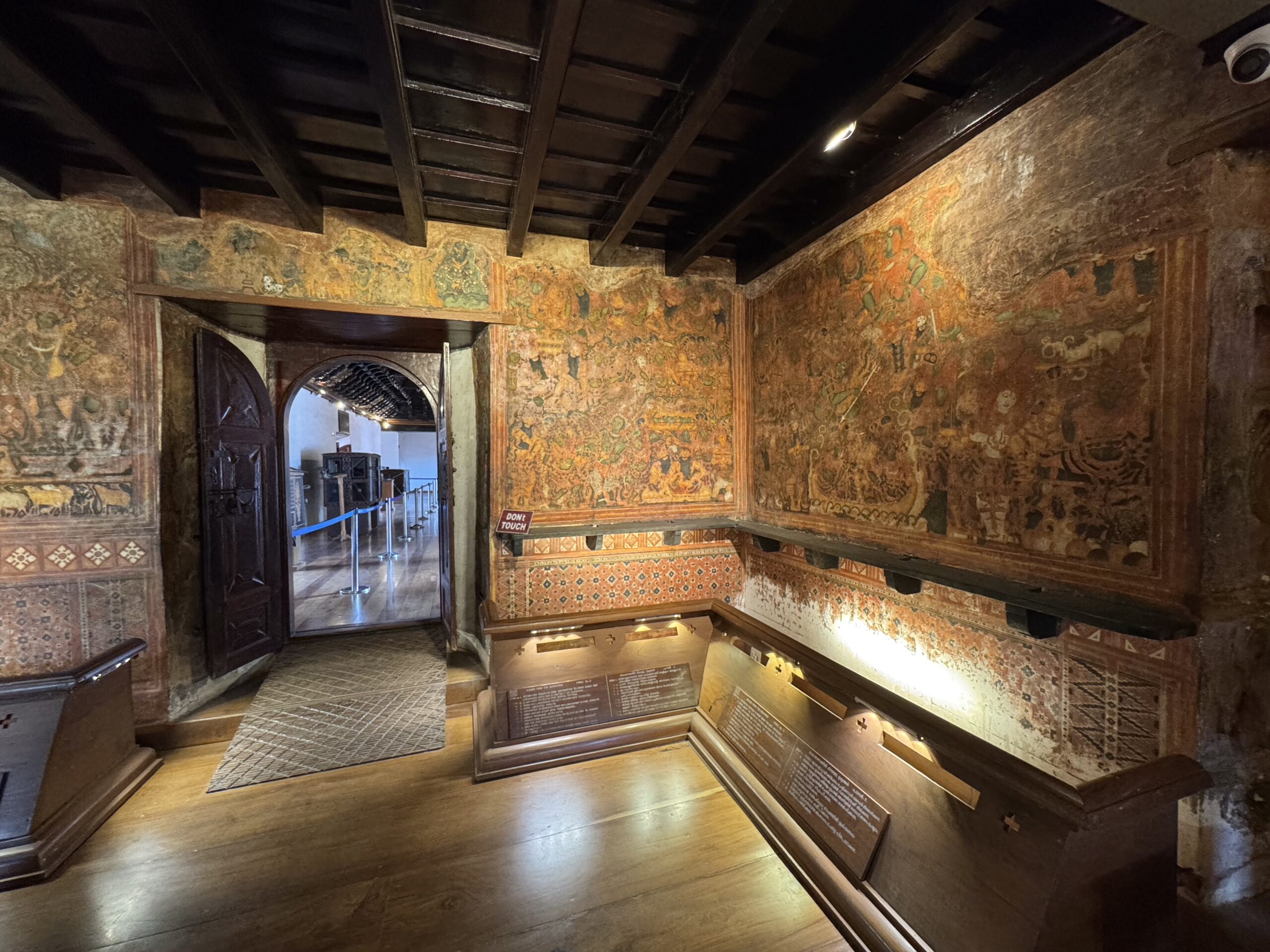 Historic room with faded wall frescoes, dark wooden ceiling beams, and glass-encased displays along the wall and railing.