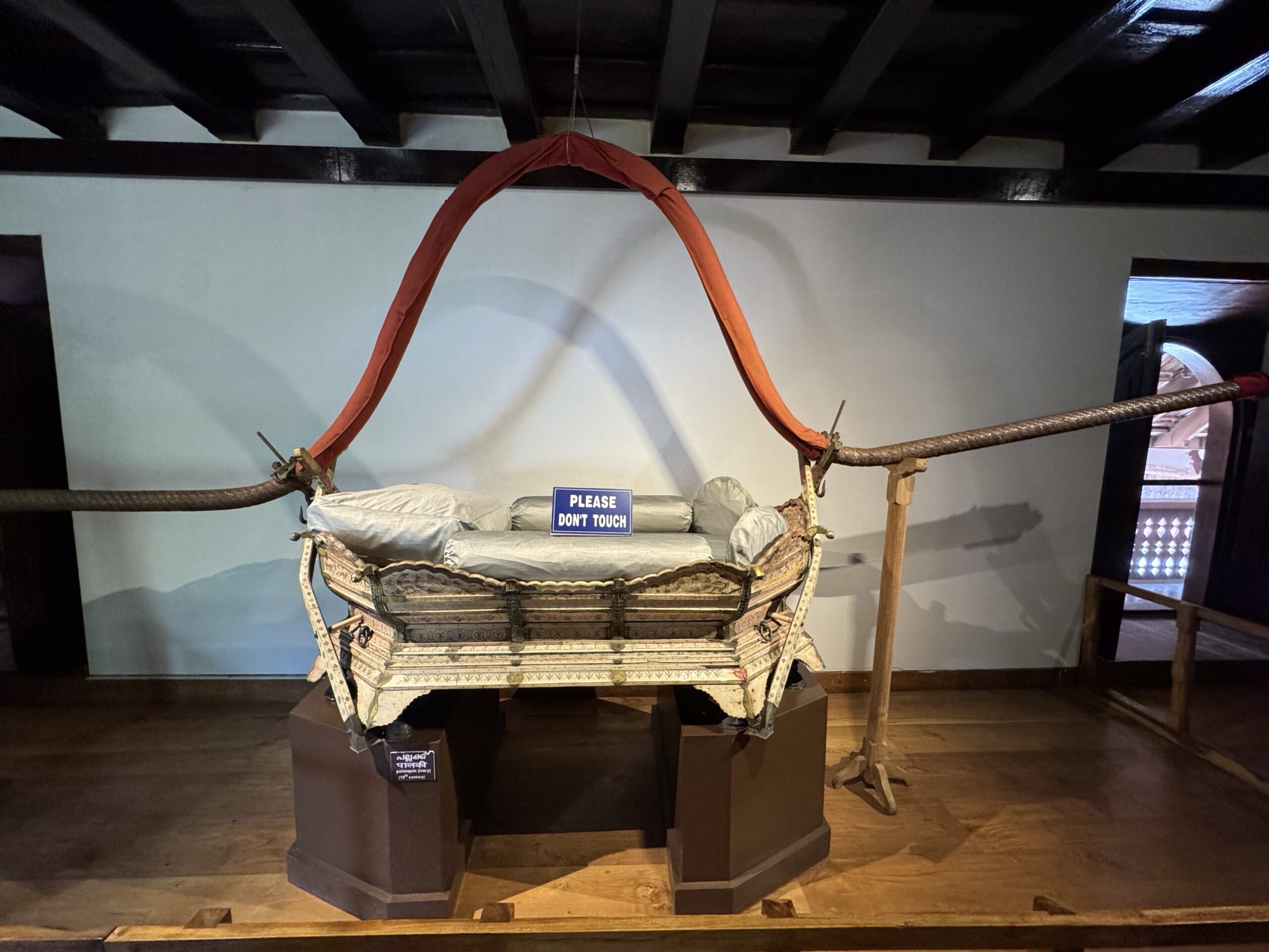 Ornate antique cradle suspended by an orange fabric arch in a museum display, with a blue 'Please Don't Touch' sign nearby.