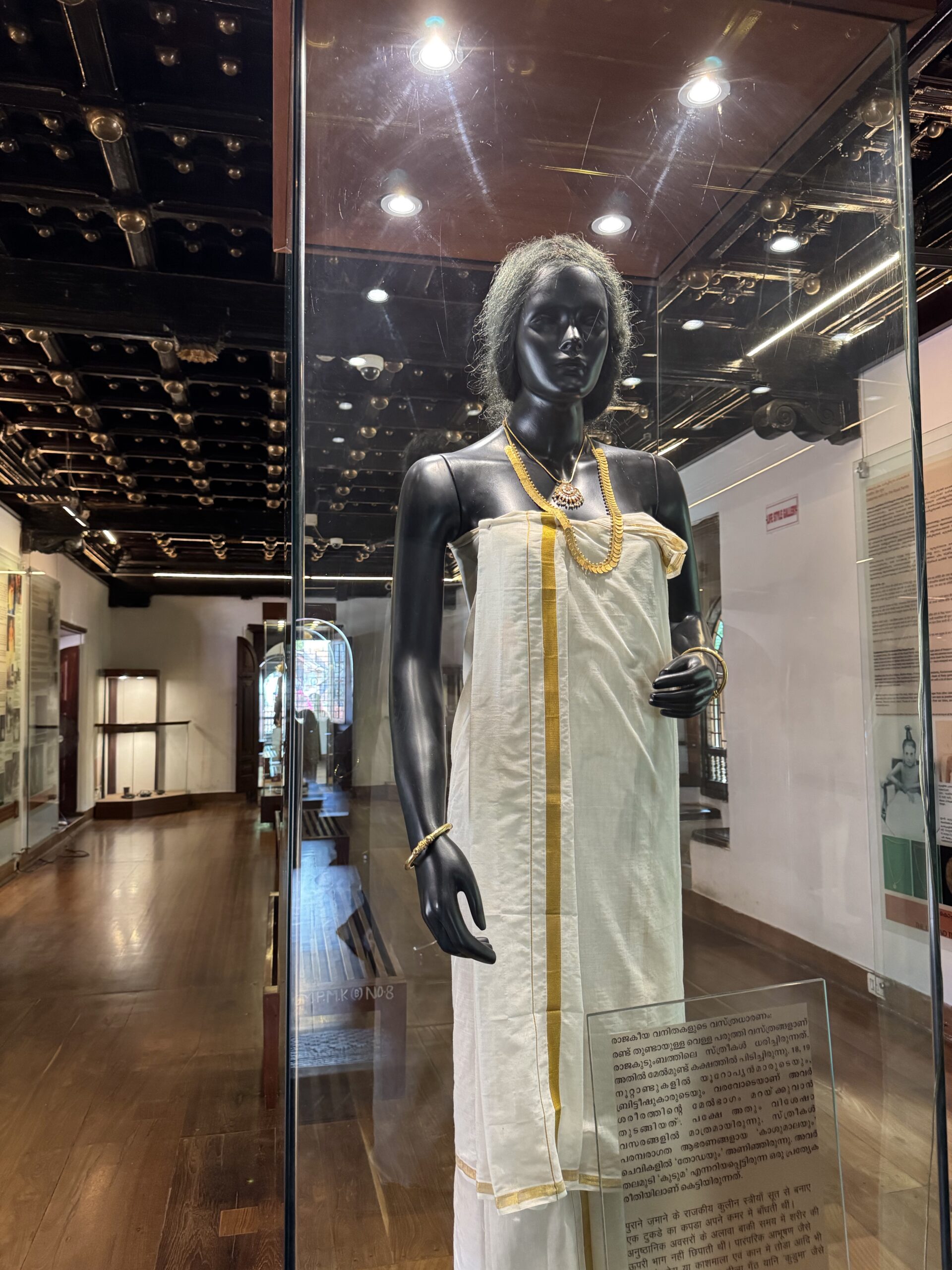 Black mannequin wearing an off-white, toga-style dress with gold trim and a layered necklace inside a glass display case in a museum.