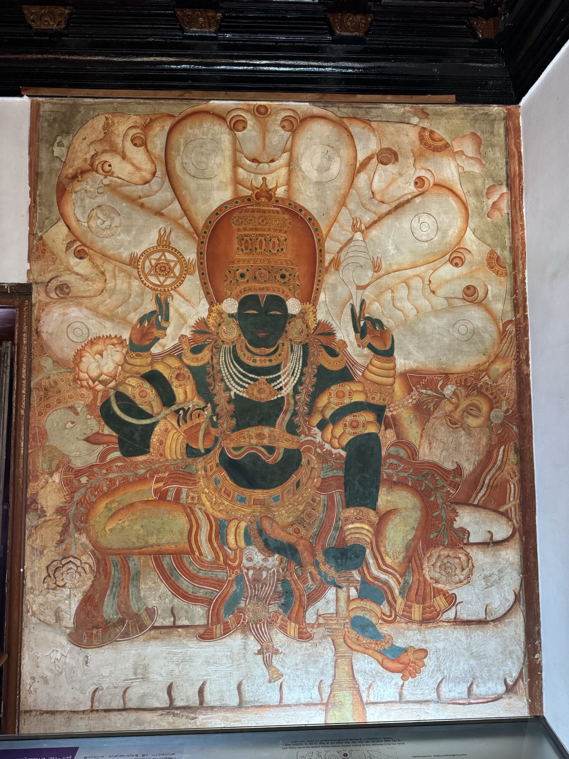 Colorful Hindu deity with an ornate crown and multiple arms, seated in a temple setting amid decorative motifs.