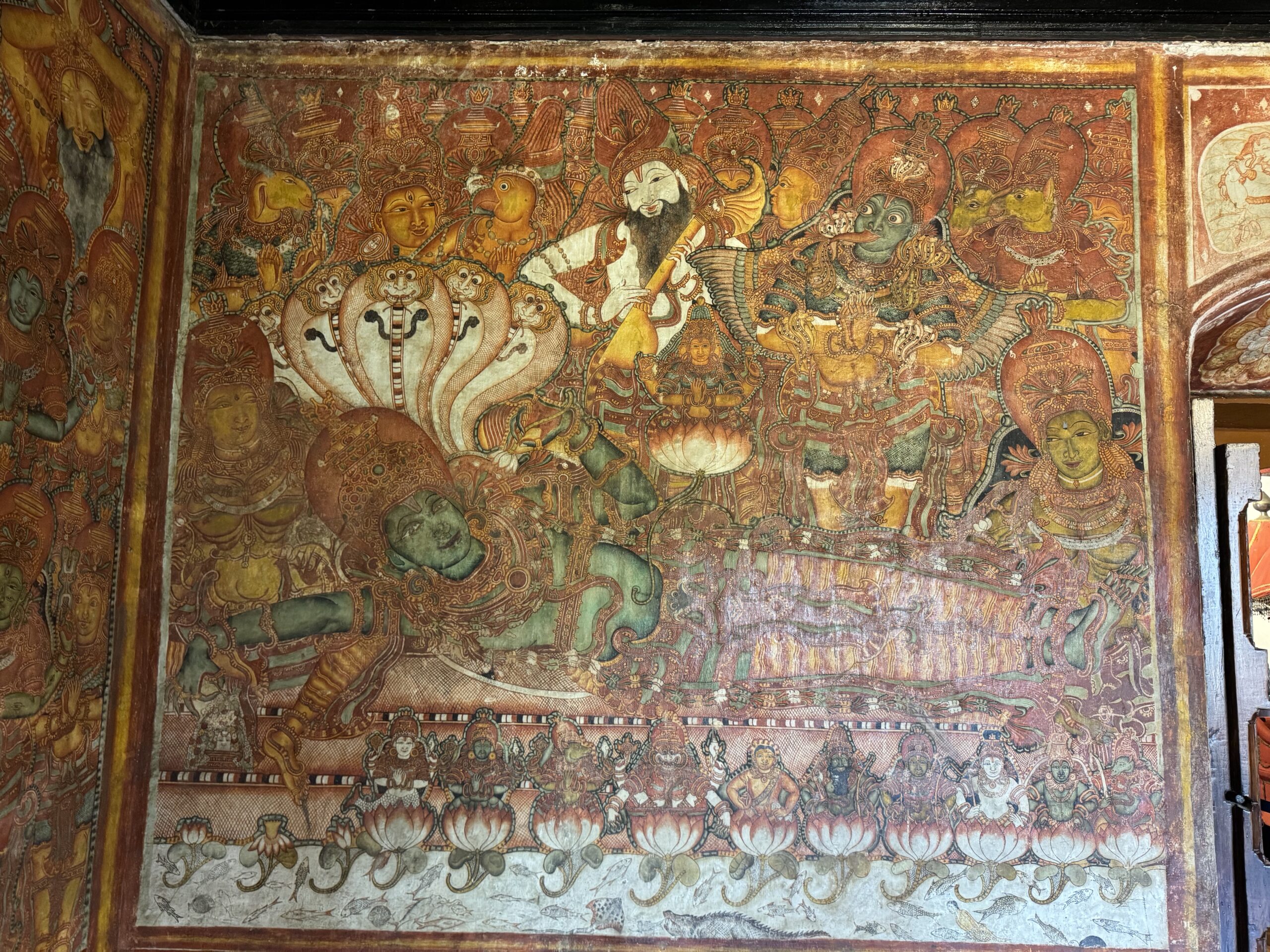 Colorful temple mural crowded with gods, demons, and serpents in a mythic scene adorned with crowns and lotus motifs at the bottom.