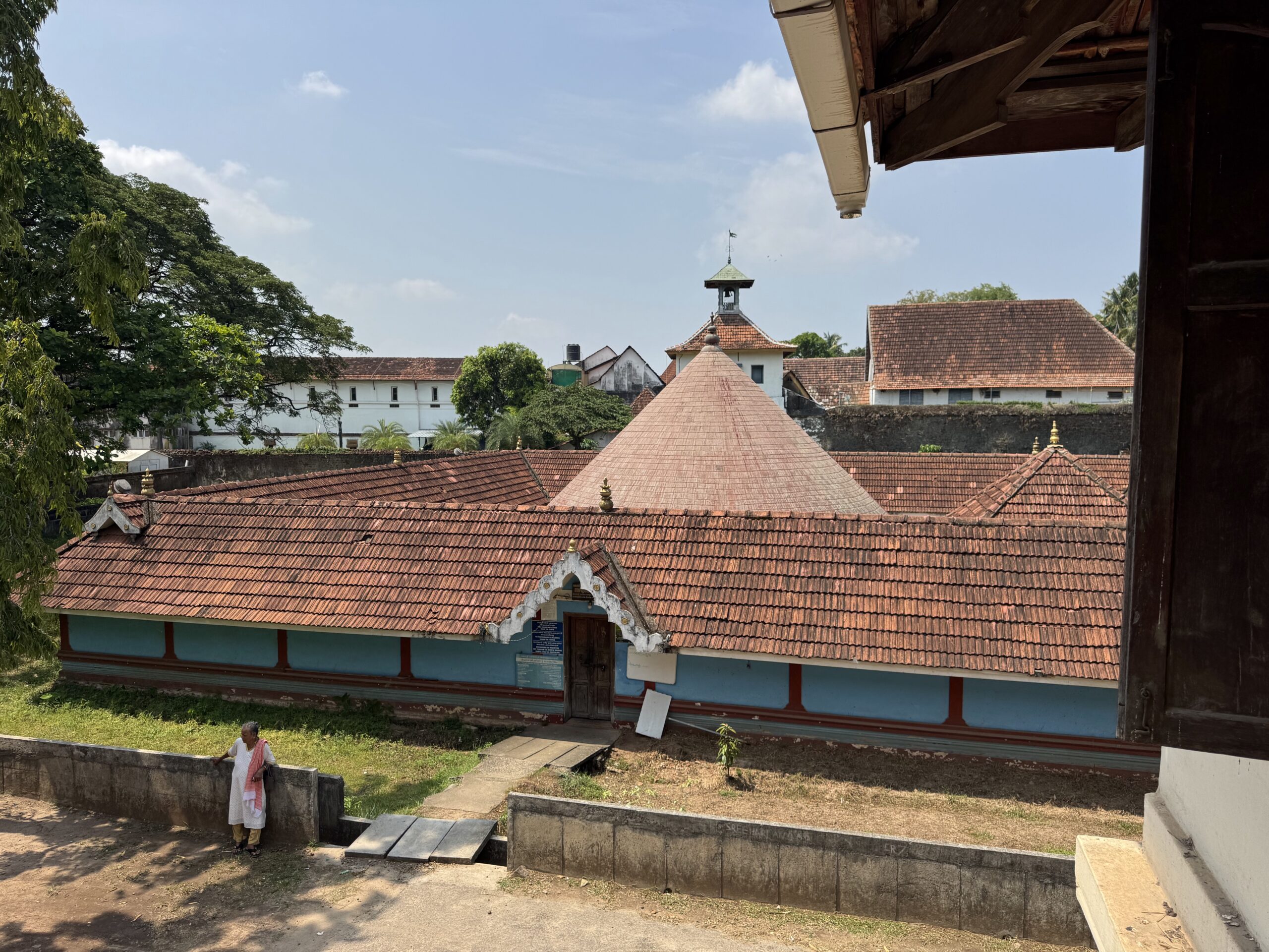 Temple complex with terracotta red tiled roofs, a small central tower, and a person walking along a low wall.