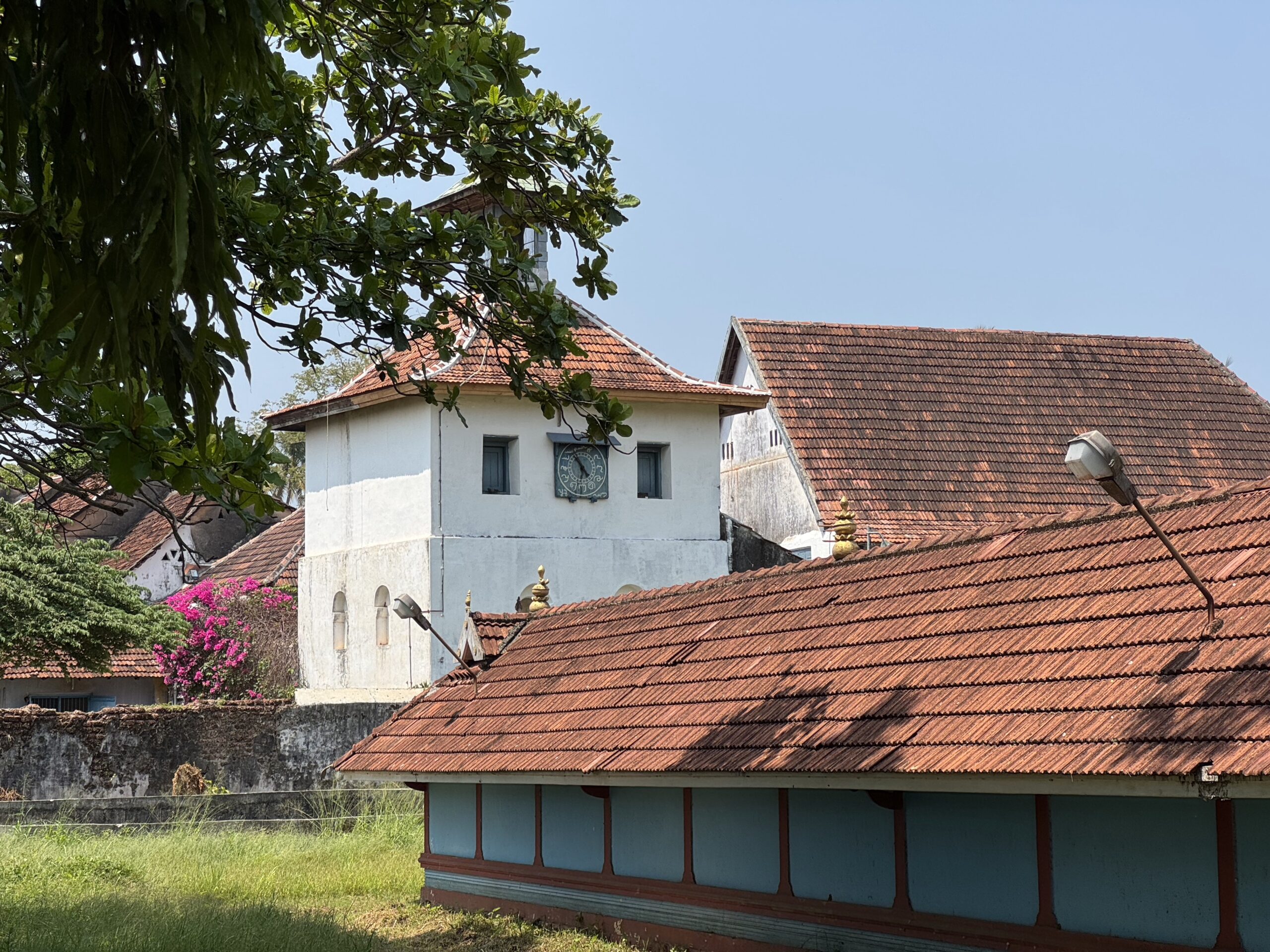 White building with red tile roof and a clock on the facade, framed by green trees against a blue sky.