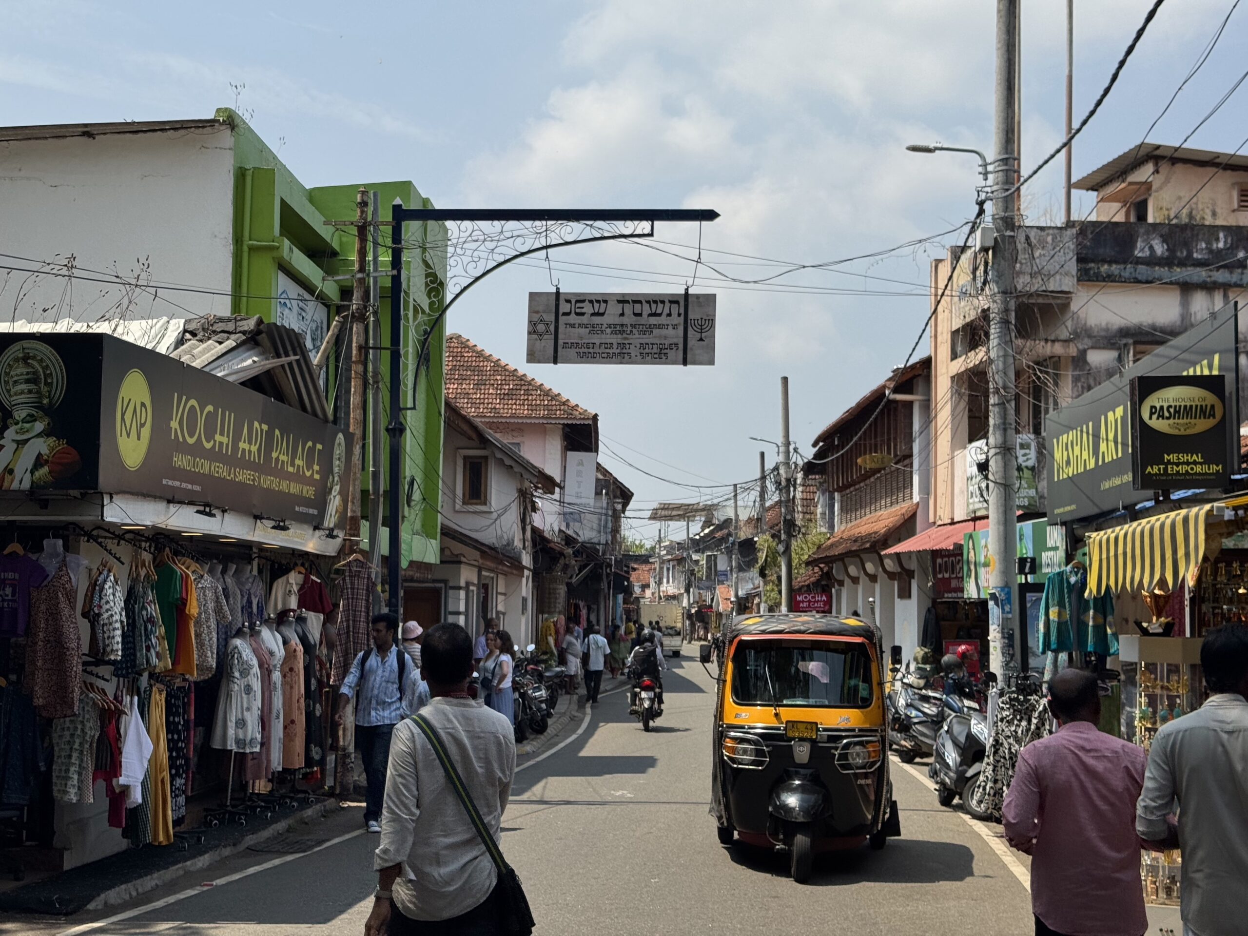 Busy street market with clothing stalls on both sides, pedestrians walking, and a yellow auto-rickshaw in the center against a bright sky.