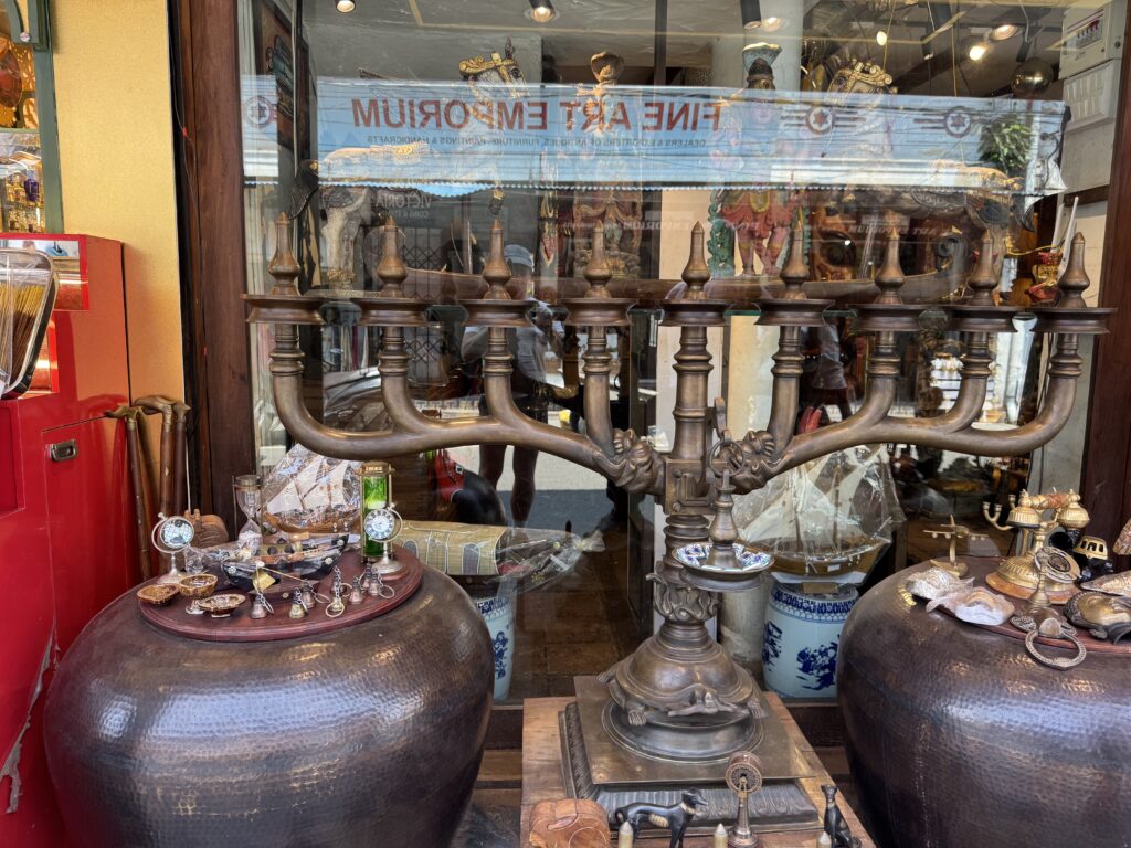Shop window display featuring large hammered metal pots, brass finials, and assorted jewelry on round tables and shelves inside the store.
