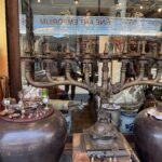 Shop window display featuring large hammered metal pots, brass finials, and assorted jewelry on round tables and shelves inside the store.