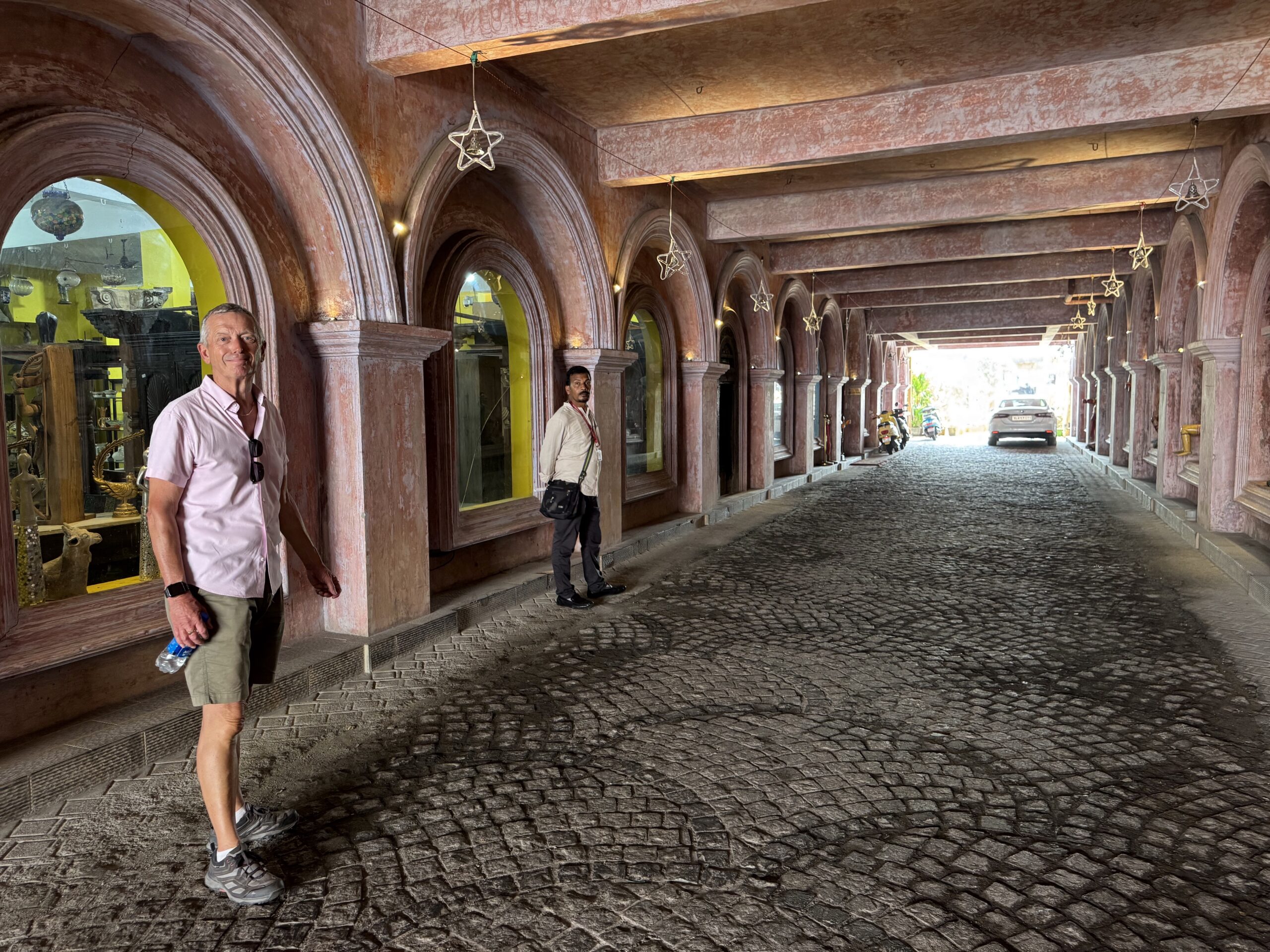 Two men stand along a long, pink-stone arcade with rounded arches and hanging star decorations on a cobblestone street at dusk/dawn light, one foreground wearing pink shirt and shorts.
