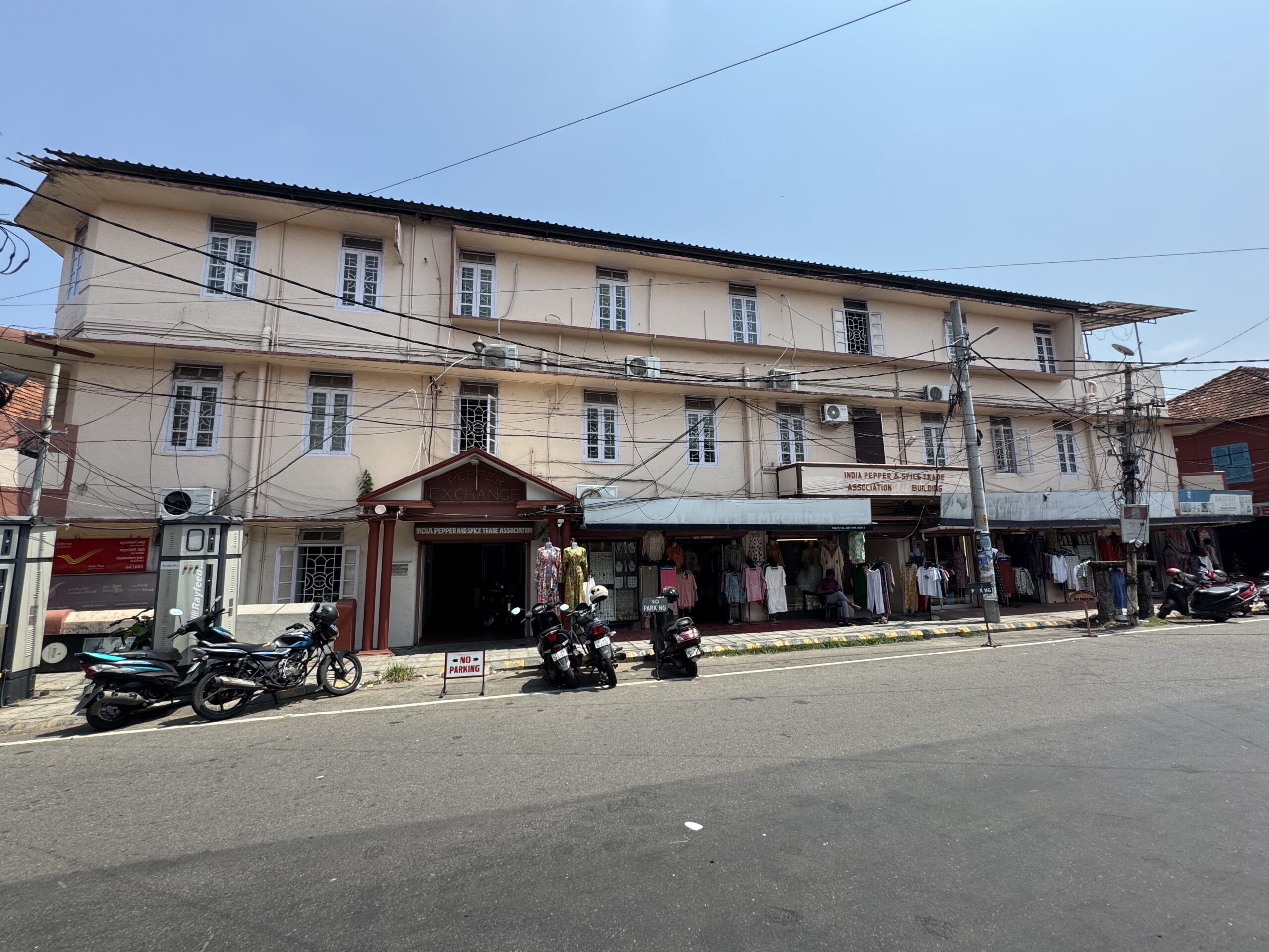 Three-story beige building with shopfronts on the ground level; clothes hanging outside and motorcycles parked along the curb under a clear blue sky