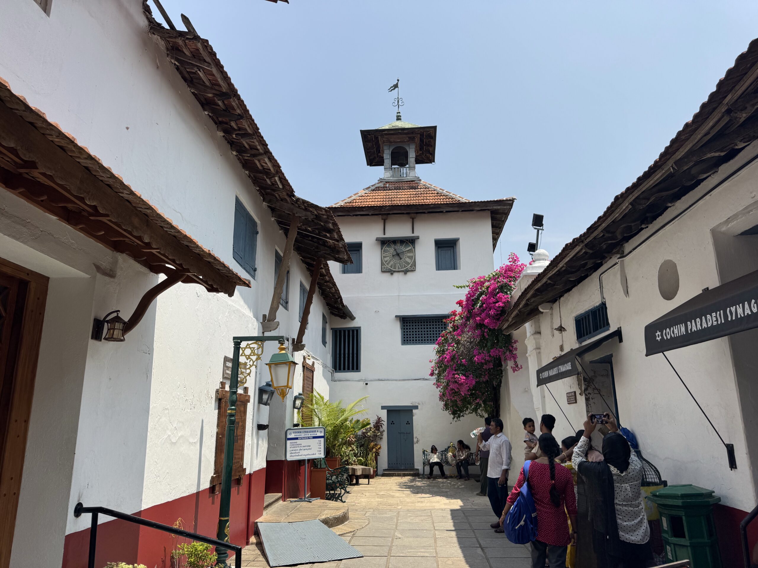 Narrow pedestrian street in a white-walled town with a clock tower in the center, flowering pink bougainvillea, blue doors, and people waiting along the storefronts during a sunny day.