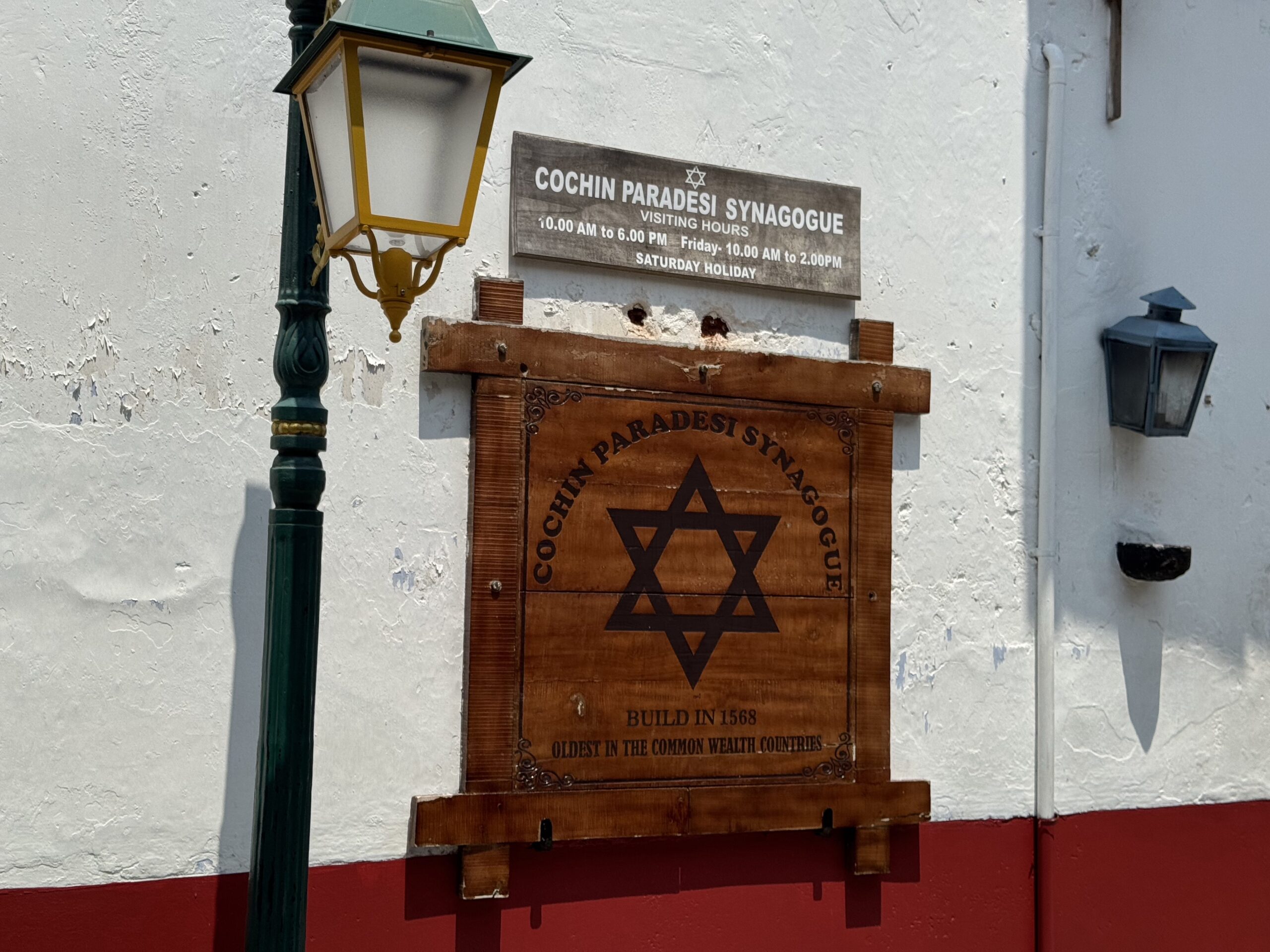 Wooden sign for Cochin Paradesi Synagogue with a Star of David on a white textured wall, beside a green street lamp (red-painted base visible).