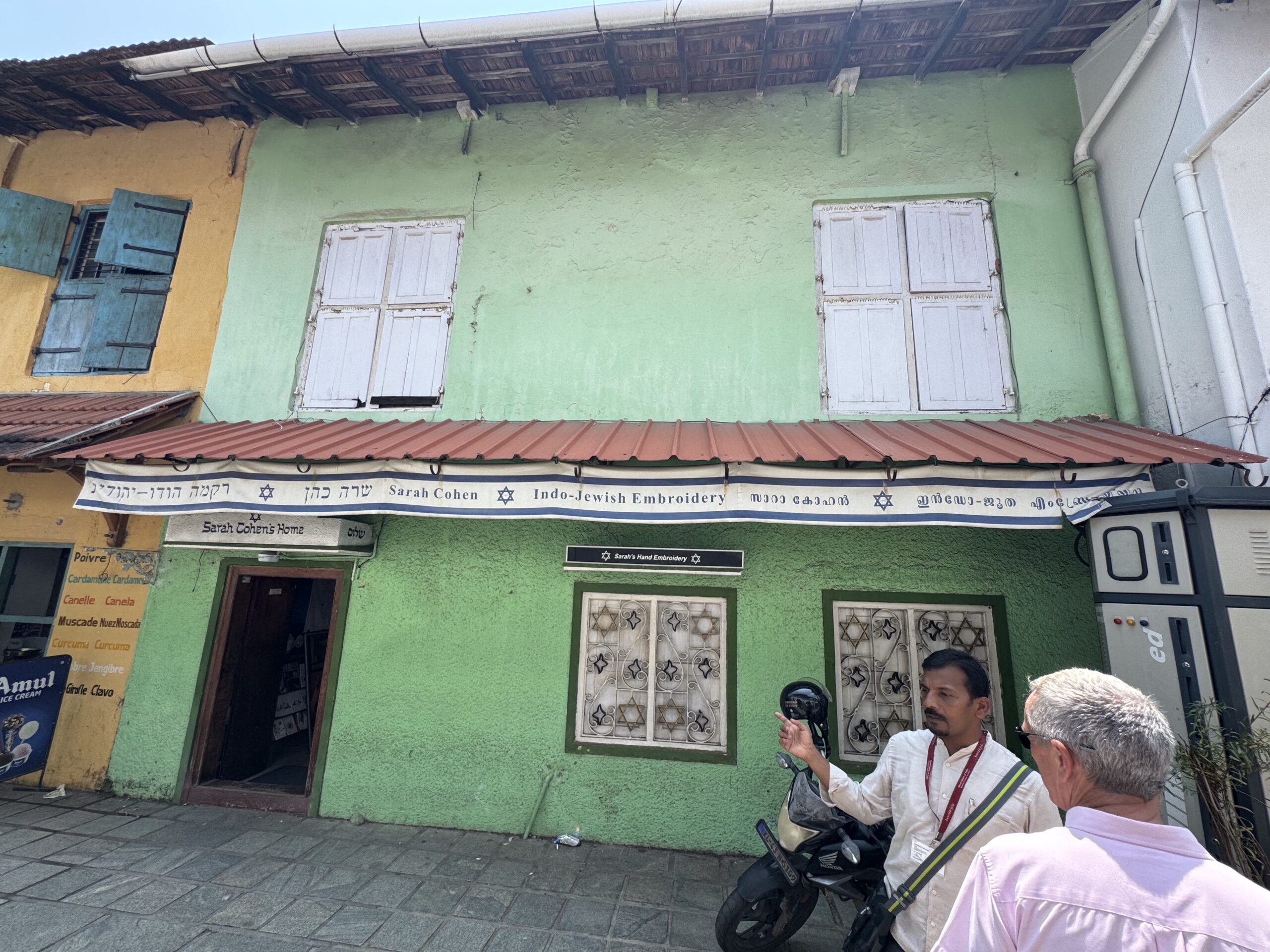 Green storefront with white shuttered windows and a banner reading 'Sarah Cohen Indo-Jewish Embroidery' across the awning.