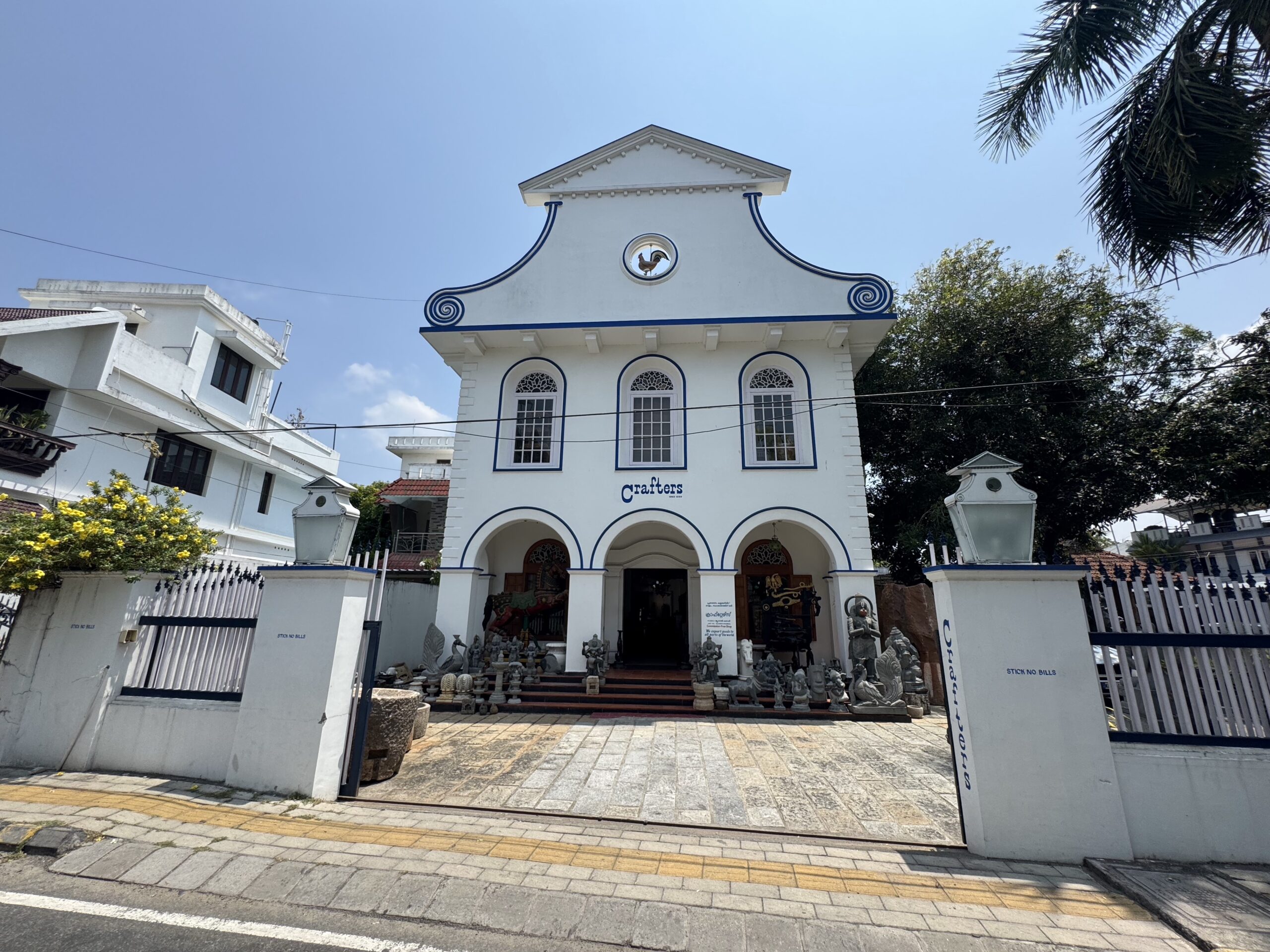 White building with blue trim and arched windows, statues at the entrance, and a sign that says 'Crafters' in front of it.