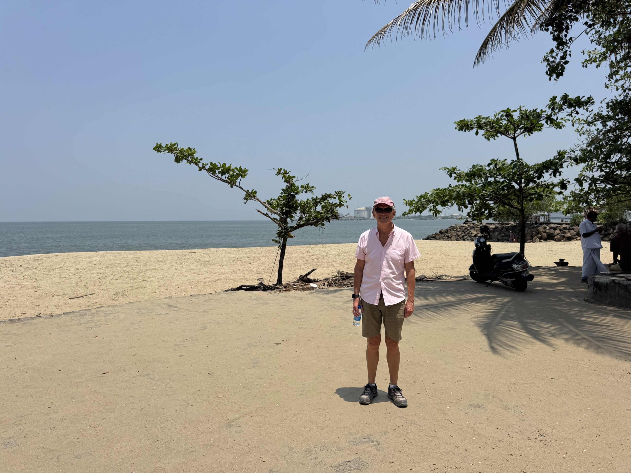 Older man in a light pink shirt and pink cap standing on a sandy beach with the sea and palm trees in the background.