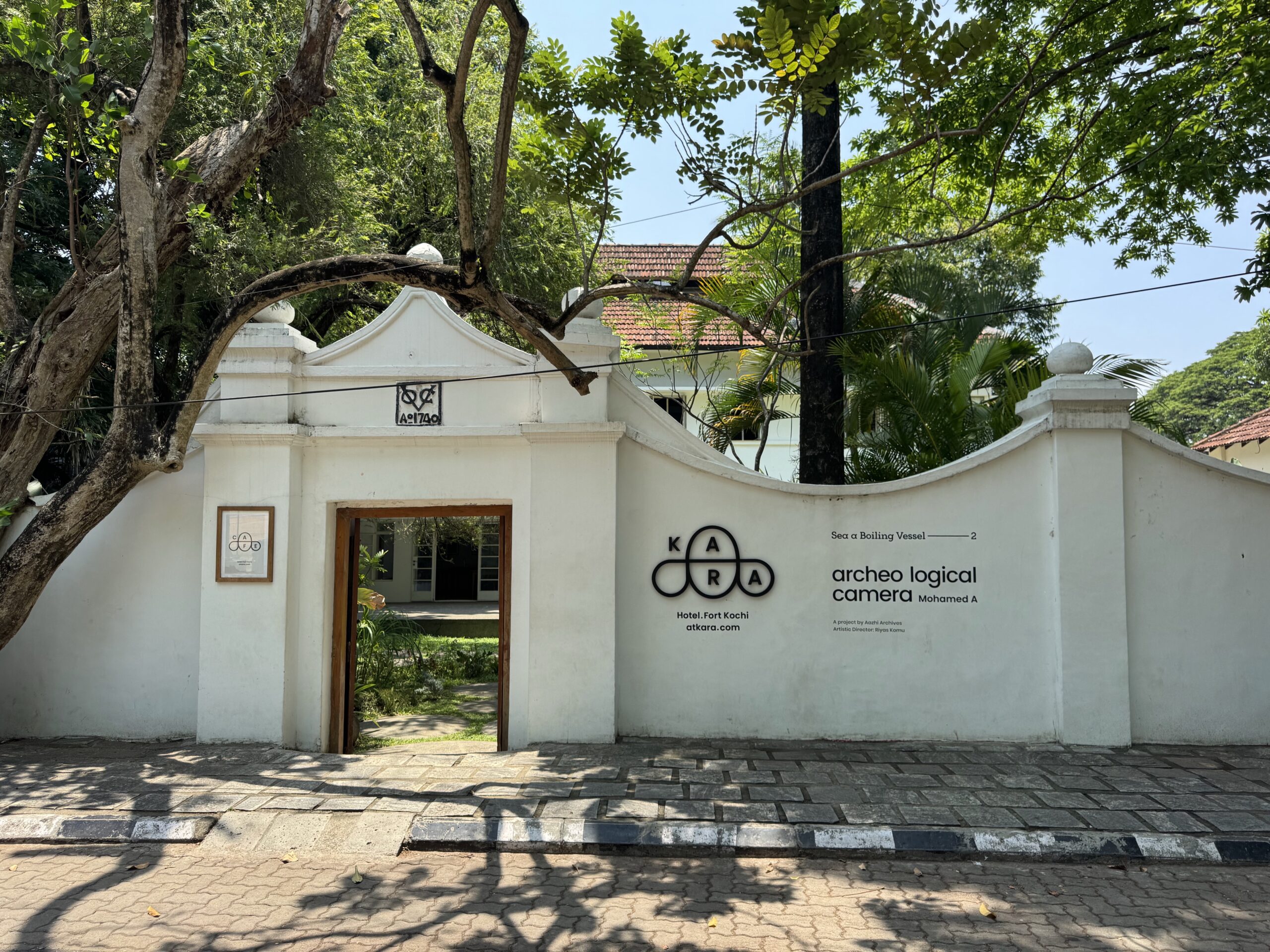Entrance to a white wall compound with an open wooden doorway, a large tree arching over the gate, and a KA logo on the wall.