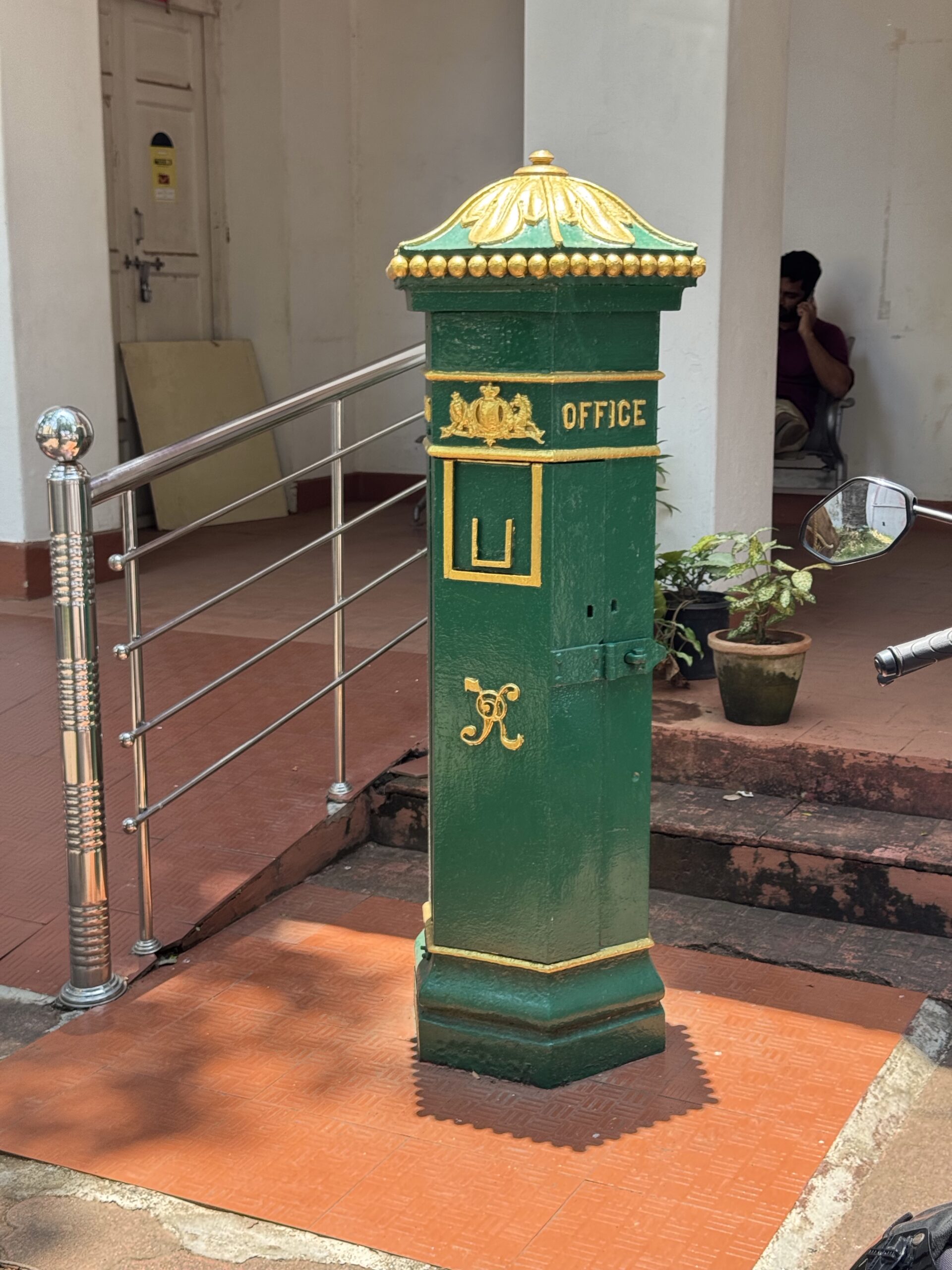 Green and gold post box with 'OFFICE' inscription and crest, standing beside a railing outdoors on a tiled path.
