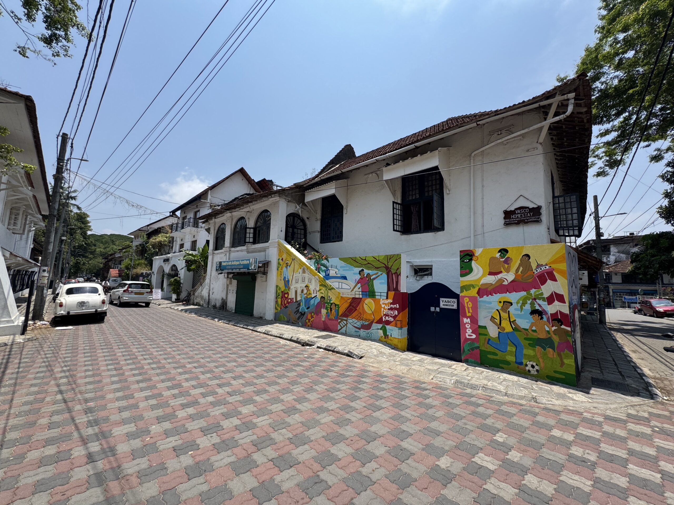 Street view with a white two-story building and a colorful mural wall along a cobblestone sidewalk