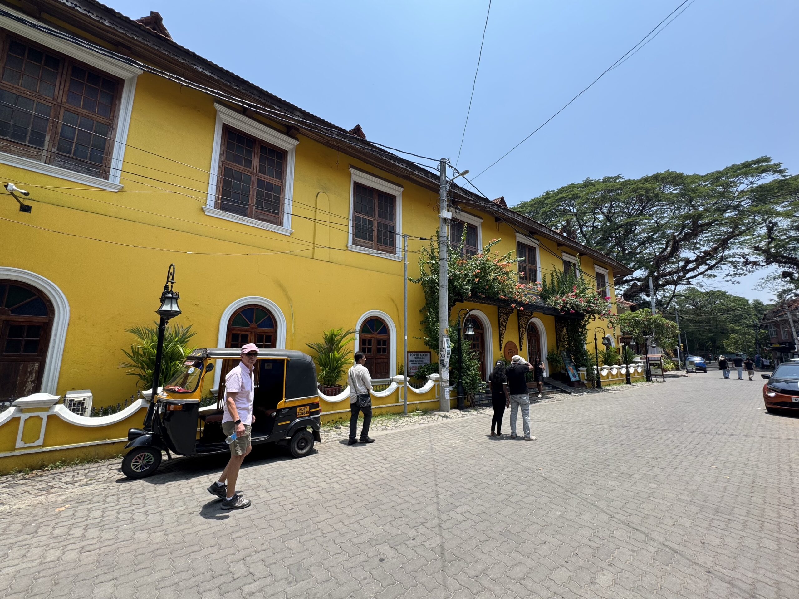 Yellow building with arched windows on a sunny street; people and a small three-wheeled vehicle nearby, power lines overhead.