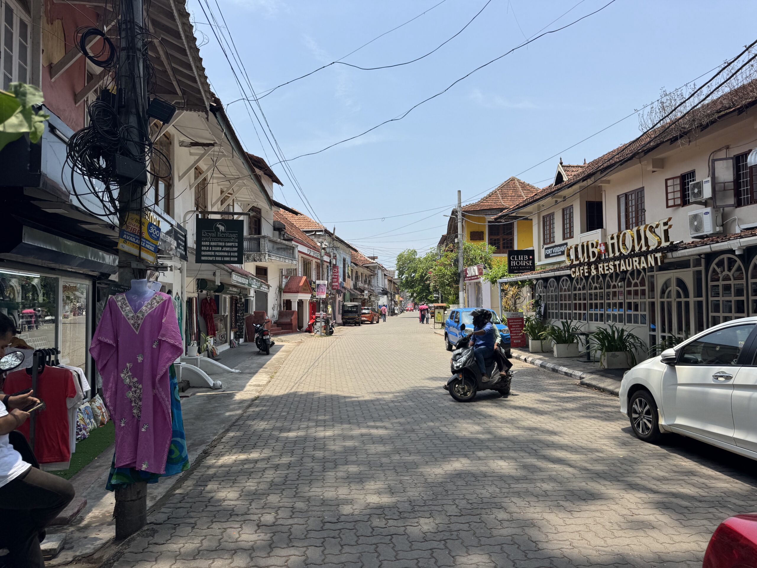 Narrow pedestrian street lined with shops and cafés, scooters parked along the curb, and a bright blue sky above with tangled overhead wires.